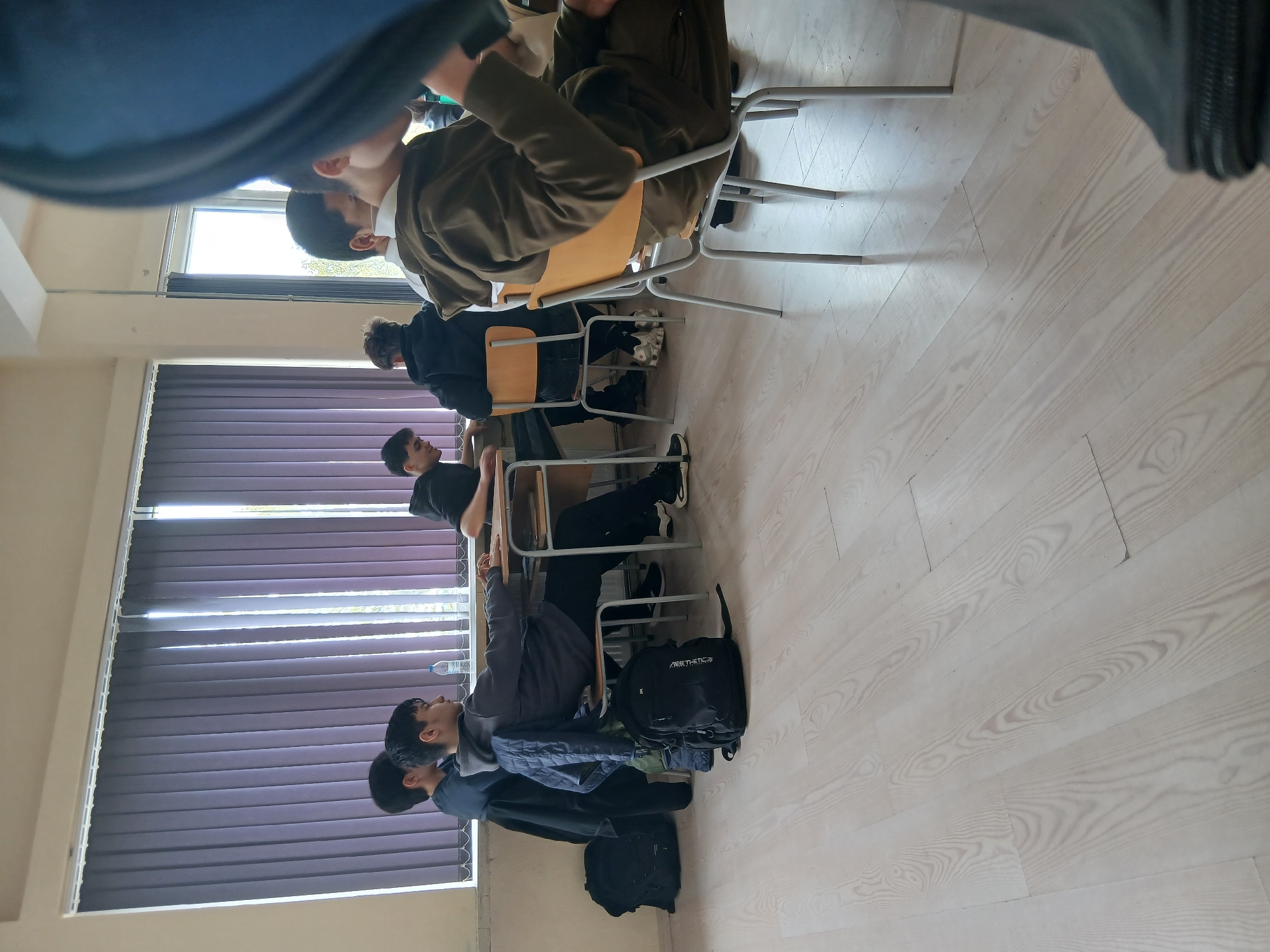 The image depicts a classroom in Varna, Bulgaria. There are four visible rows of students sitting in chairs, presumably attending a lesson. Each student is facing the same direction, with their desks in front of them. Some have bags placed under their chairs. A set of blinds covers a window along one side of the room. The room has light-colored wooden floors and pale walls.