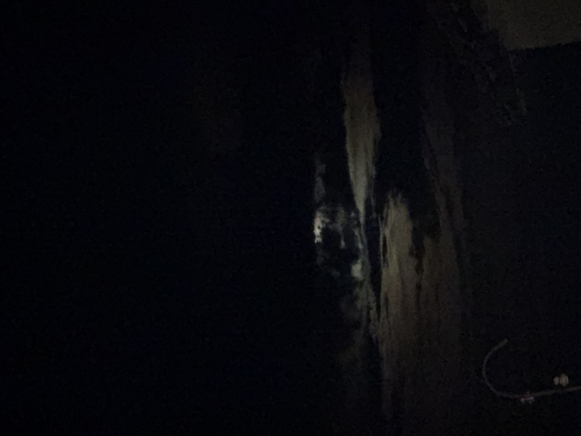 The image shows a dark sky with scattered clouds illuminated by moonlight. The clouds appear to be moving, creating a dynamic texture across the sky. In the lower left corner, a curved object, possibly a streetlamp or part of a fence, is visible. The rest of the scene is dark and indistinct, suggesting it is nighttime. There are no people or discernible structures in the image. The scene is generic and does not contain any specific visual elements that would indicate a location in Pavel Banya, Bulgaria, or any other city.