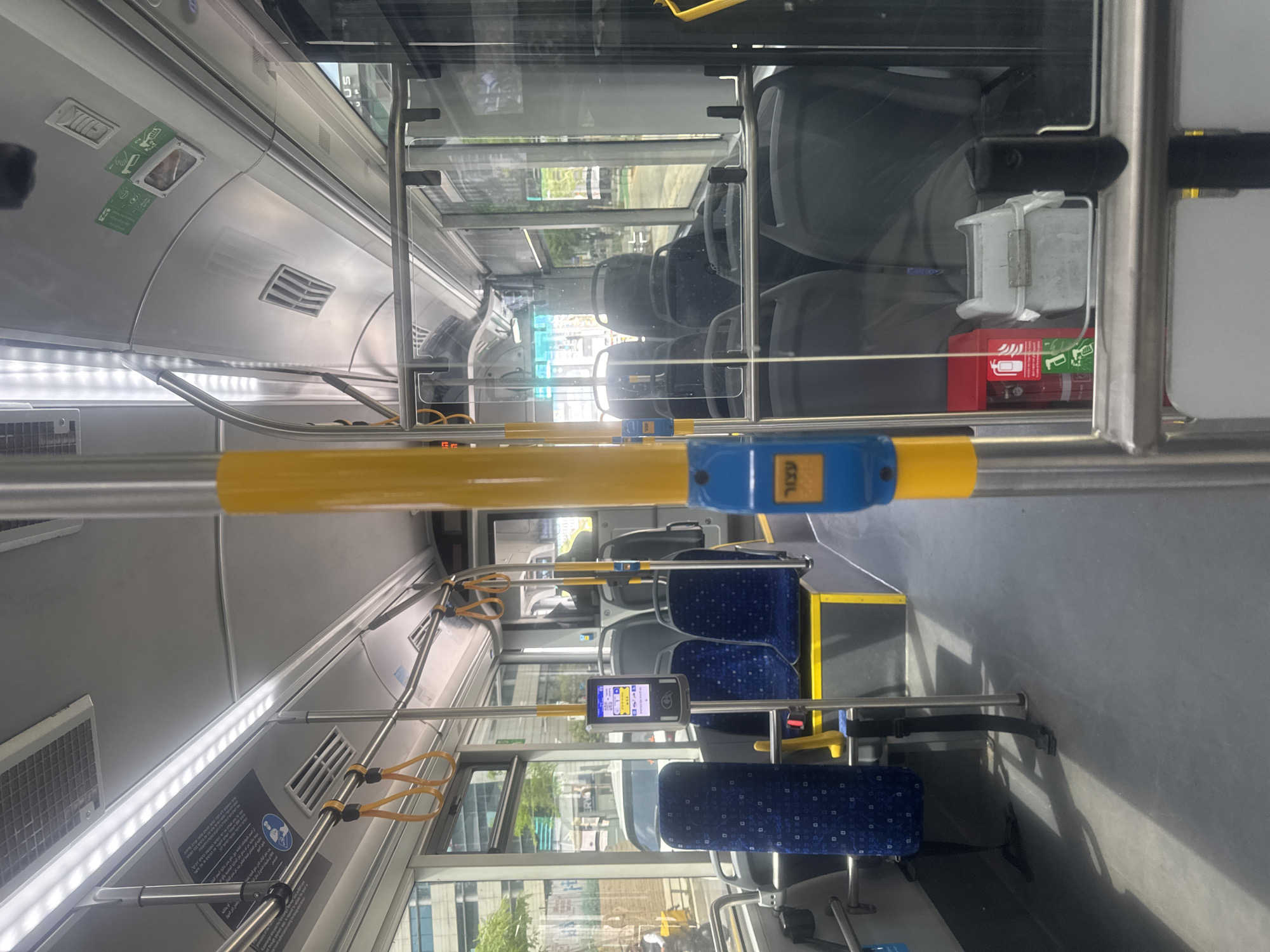 The image shows the interior of a public bus, likely in Tel Aviv, Israel. The bus appears to be nearly empty. Rows of blue and gray upholstered seats are visible. Yellow handrails and blue stop-request buttons are prominent. A fare payment machine with a screen displays information. A glass barrier separates the front section. Overhead, lighting panels are visible, along with bus safety and information signage. A fire extinguisher is mounted near the front of the bus behind the glass barrier. Through the windows, urban buildings and greenery can be seen, suggesting a city street scene.