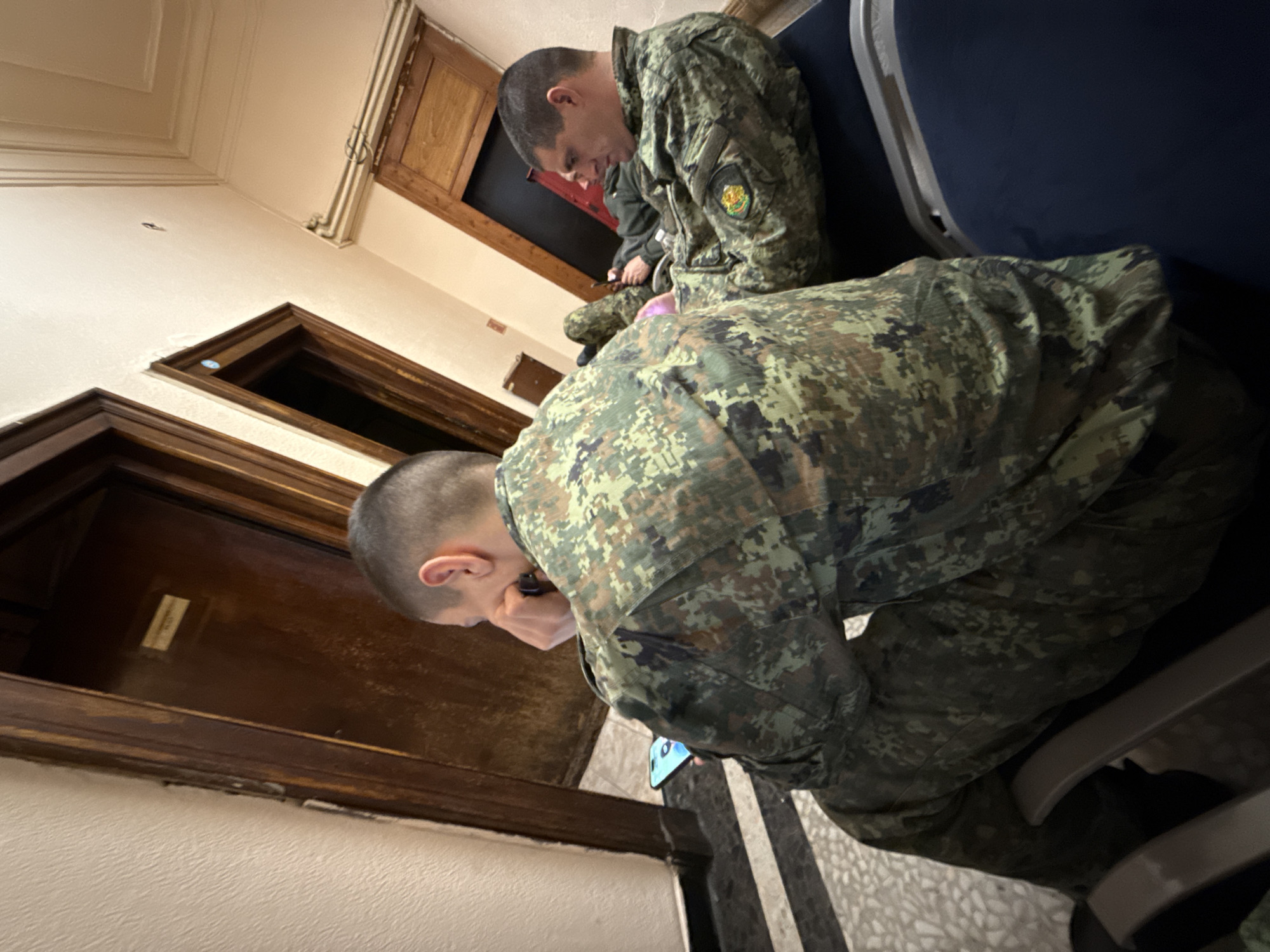 Two men in military uniforms are seated in an interior location. They are focused on their electronic devices. The uniforms have camouflage patterns. The location appears to be a hallway with wooden doors and possibly seats along a wall. The presence of military personnel suggests an institutional setting. There is no definitive information in the image to pinpoint the location in Sofia, Bulgaria.