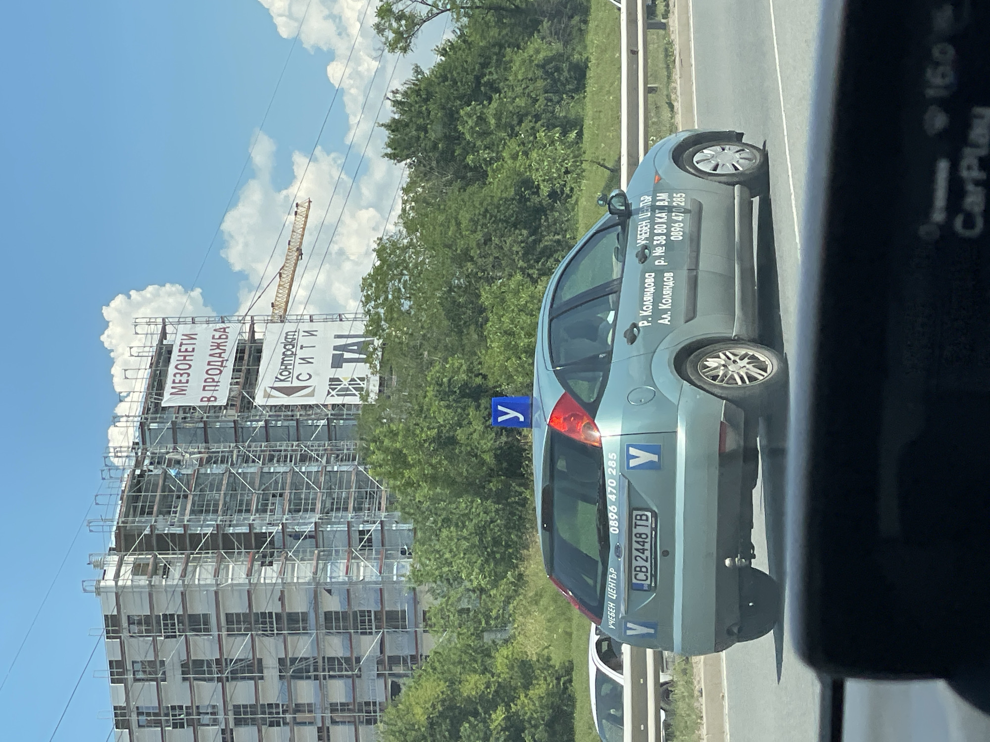This image captures a bright, sunny day in Sofia, Bulgaria. The scene is dominated by a building under construction, encased in scaffolding and adorned with banners. One prominent banner displays text in Bulgarian, including "МЕЗОНЕТИ В ПРОДАЖБА" (Maisonettes for Sale) and "КОНТРОЛ" (Control), with additional text likely related to the building project.
In the foreground, a light teal car is parked on the side of a road, also displaying Bulgarian text. The text on the car identifies it as belonging to a "УЧЕБЕН ЦЕНТЪР" (Training Center) and includes contact information: "Р. Коляндова" (a name), "Ал. Коляндов" (another name), a street address "Р. № 38 80 КАТ. В.М" (Street No. 38, Block 80, Section B.M), and a phone number "0896 470 285". The car also features a license plate "CB 2448 TB" and several blue square signs with a white "Y" symbol, likely indicating a driving school vehicle.
The sky is a clear blue with fluffy white clouds, and lush green trees line the background, suggesting a warm season. The presence of a construction crane near the building further reinforces the idea of ongoing development. The image appears to be taken from within another vehicle, as indicated by a dark, reflective surface on the right side of the frame, possibly a car window or dashboard with "CarPlay" visible. The overall impression is a snapshot of urban life with construction, commerce, and everyday activities.