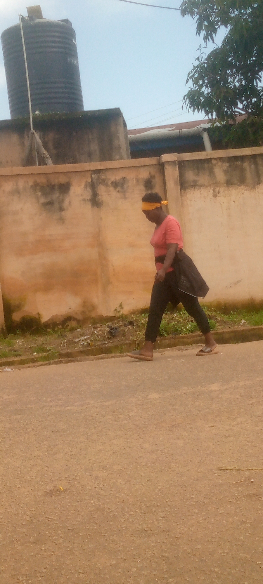 A woman with a yellow bandana around her head is walking on a paved surface. She is wearing a pink t-shirt, black pants, and brown sandals. A dark jacket is draped over her left shoulder. The woman is walking from left to right in the frame. Behind her is a textured, light-colored wall. In the background, a large black water tank is visible on top of a structure, along with trees and the sky. The scene appears to be outdoors, possibly in a residential or commercial area of Jos, Nigeria, given the architecture and vegetation. The lighting suggests it is daytime.