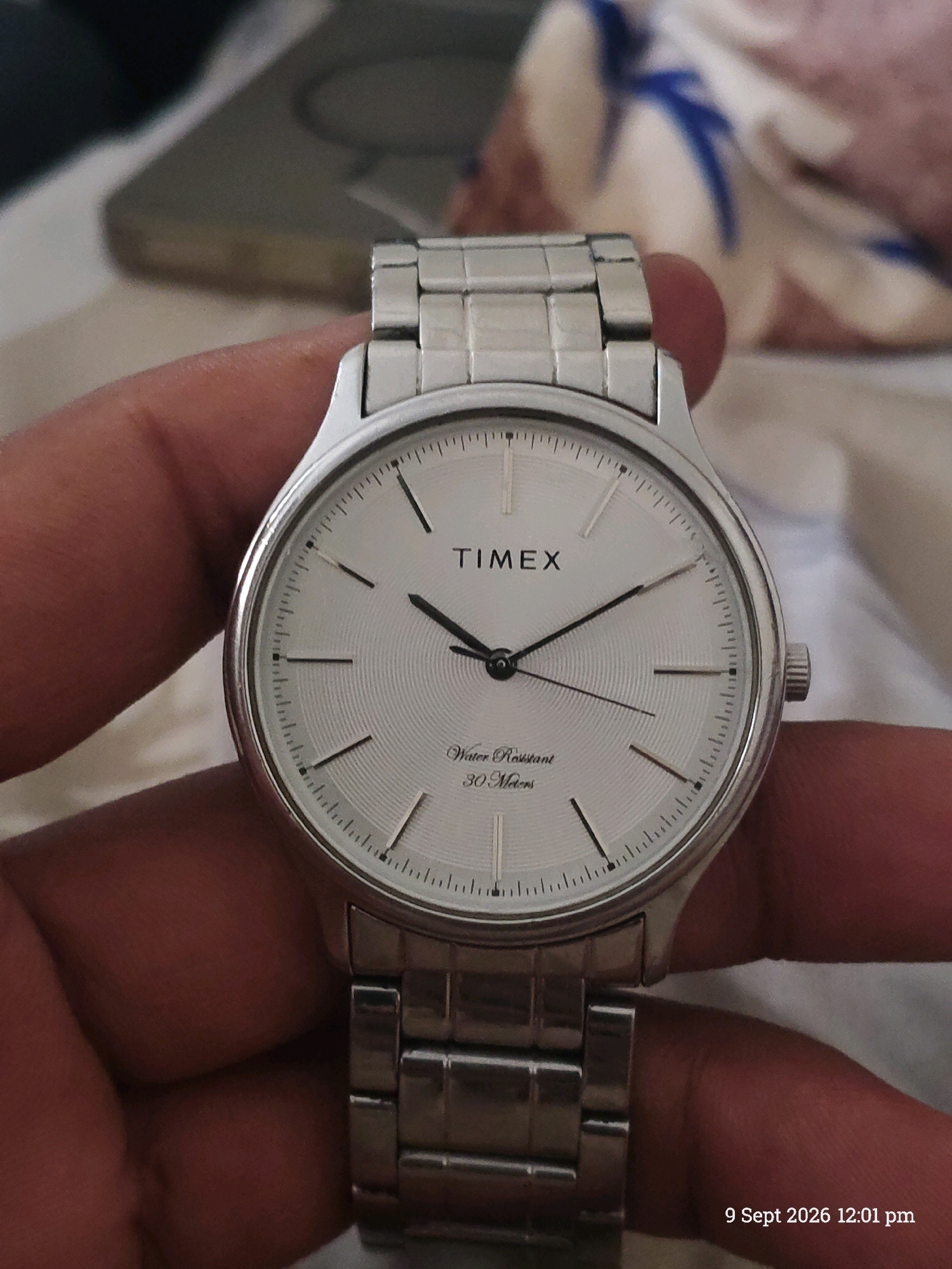 A close-up shot shows a person's hand holding a silver Timex watch with a white dial. The watch has a metal band and black hands. The background is blurred and appears to be a bed with a patterned blanket. A timestamp in the lower right corner reads "9 Sept 2026 12:01 pm". The image does not provide any information about the location within the city of Indore, India.