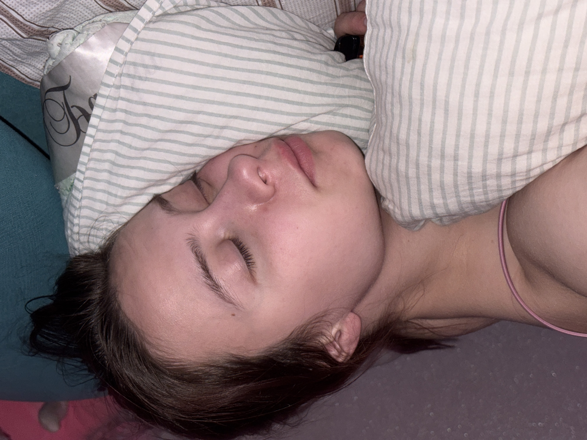 A person is lying in bed. The person has fair skin, dark hair, and closed eyes. They are under a pillow with light green and white stripes. Part of a silk pillowcase with a dark design is visible. A pink bra strap is visible on the right side of the image. The background appears to be a bedroom setting. The location (Прикамский, Russia) is not discernible from the image.