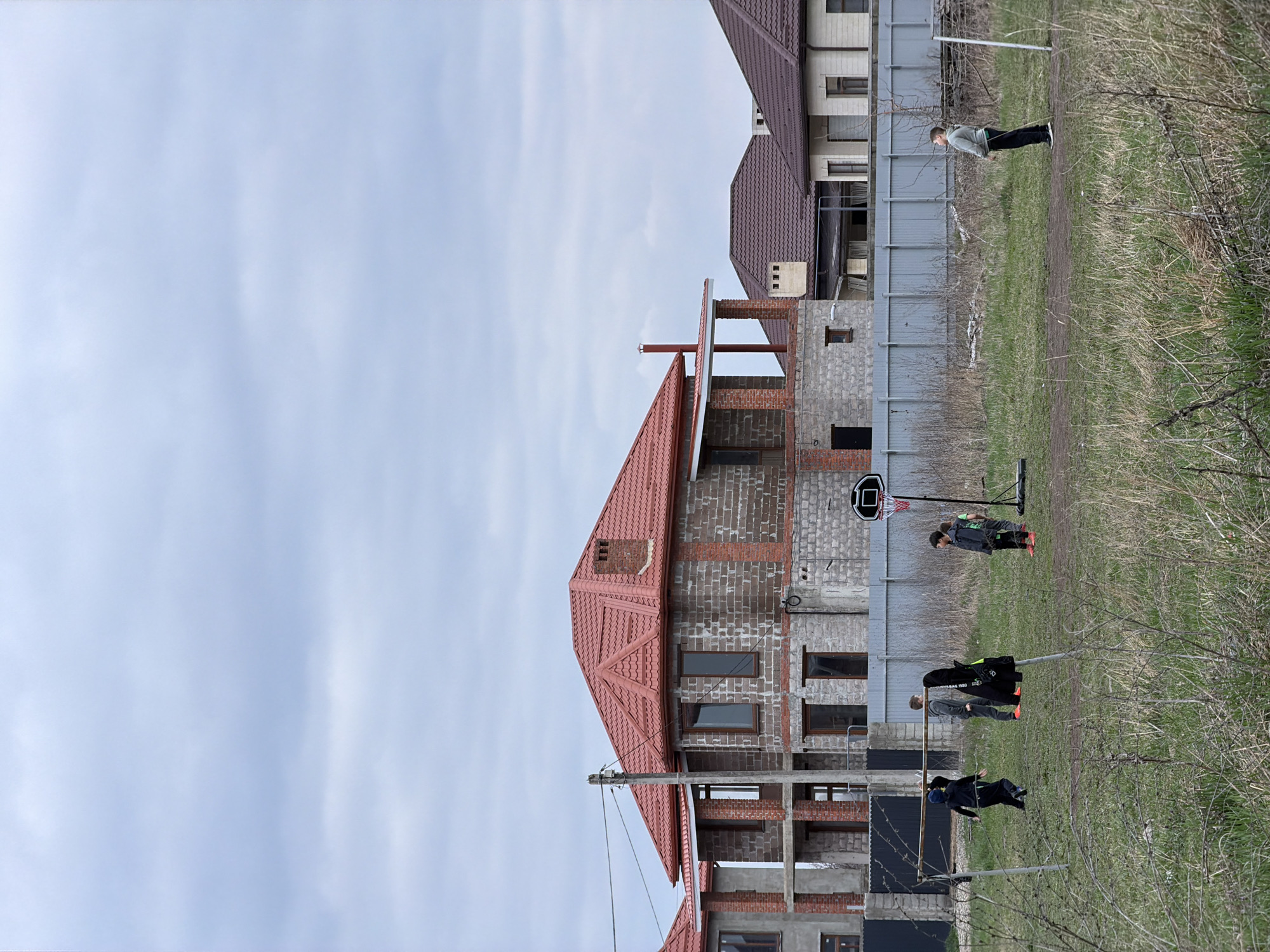 The image depicts an outdoor scene in Rossiyskiy, Russia, under an overcast sky. The foreground is an unkempt field of dry and green grass with dirt patches. Five individuals, appearing to be male children or young teenagers, are present in the field. On the left, three are grouped near a makeshift goalpost constructed from two vertical poles and a horizontal crossbar. One child, dressed in dark attire, moves towards the left; another, in a grey tracksuit, stands facing right. A dark jacket hangs from one of the goalpost's vertical supports. Centrally, a portable basketball hoop with a black backboard and red rim is positioned. Another child, wearing dark clothing and red footwear, stands near this hoop. On the far right, a fifth child, clad in a light grey hoodie and black pants, stands facing right.
A corrugated grey metal fence separates the field from the background residential area. Behind the fence, a two-story house under construction is prominently featured, characterized by a red-tiled roof, exposed brickwork, and concrete block walls. Dark-framed windows are installed in several openings. A utility pole with multiple overhead wires stands in front of this construction. To the right of the partially built house, several completed residential houses with dark brown tiled roofs and lighter facades are visible, suggesting a developing suburban environment.