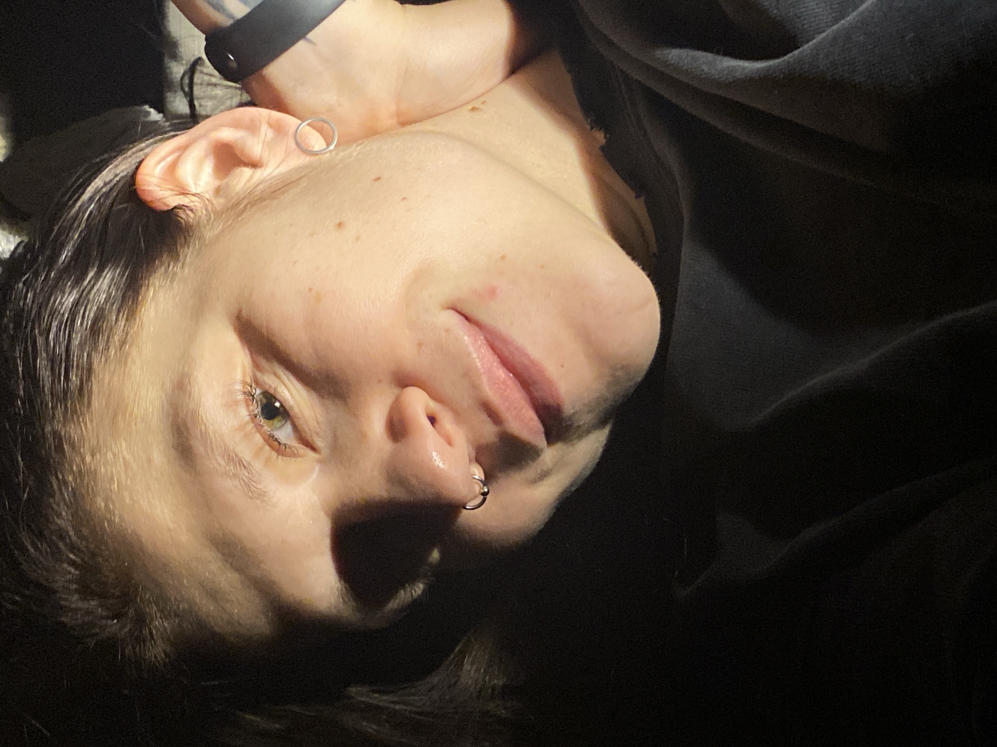An eye-level portrait shows a person lying down, with their head tilted, likely looking upwards or towards the camera. The subject's face occupies the left side of the frame, angled, with strong directional lighting highlighting features such as the forehead, nose, and lips, while casting significant shadows on the lower part of the face and neck.
The person has dark hair, which appears somewhat damp or styled close to the head. Their left eye is open, revealing a green or hazel iris, and eyelashes are visible. A silver hoop earring is worn in the cartilage of the visible ear, and another silver hoop with a small bead pierces the left nostril. Freckles or small moles are visible across the skin, particularly on the cheek and chin. A small reddish blemish is present near the corner of the mouth. A dark watch or wristband is visible on the wrist, partially obscured by the head.
The person's expression appears neutral or relaxed, with a slight openness to the lips. The background and right side of the image are dominated by dark fabric, likely clothing such as a hoodie or sweater, creating a soft, shadowed backdrop. The overall lighting suggests an indoor setting or evening light, with a single, strong light source from above and to the left of the subject. There is no visible text in the image. The scene takes place in Chaikovsky, Russia.