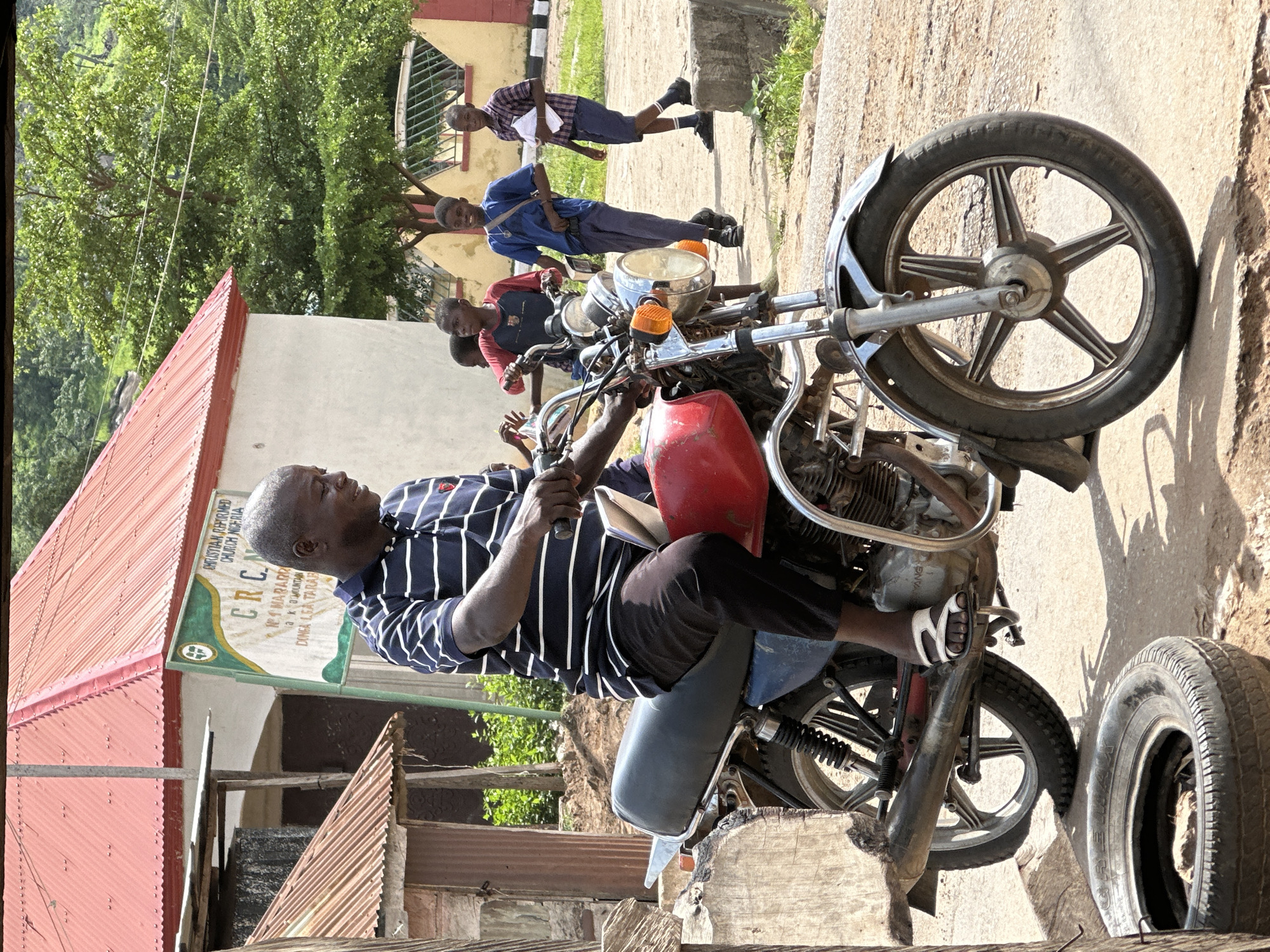 In Mararaba, Nigeria, a Black man in a blue and white striped polo shirt and dark pants rides a red motorcycle. He is looking to his right and has a slight smile. Several young Black boys in casual clothing stand behind him. One boy in a blue shirt and dark pants is looking at the camera and smiling. Another boy in a red shirt is looking at the man. A third boy in a plaid shirt and dark shorts is walking towards the right side of the image, holding a white paper. The motorcycle has chrome accents and a black seat. Behind the man and boys is a building with a red roof and a sign that reads "CHRISTIAN REFORMED CHURCH NIGERIA" and "CRC". The environment appears to be an outdoor area with a dirt path and some vegetation. There is also a tire lying on the ground in the foreground.