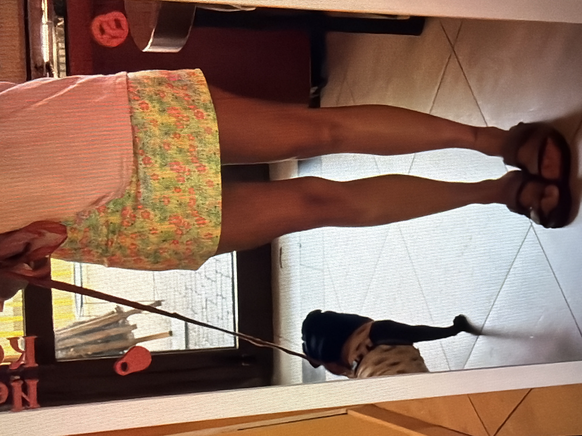 This image shows a person's legs from the knee down, wearing a light pink top and a floral skirt. The person is wearing brown sandals. They are holding a leash attached to a dog, which is partially visible in the lower right of the frame. The dog is lying down. In the background, there is a window with what appears to be a stack of rolled-up mats or similar items. There is also some red lettering on a dark surface to the left of the window. A red pretzel-shaped object is visible on a dark surface in the upper left of the image. The scene appears to be indoors, possibly a home or a shop, given the floor tiling and the items in the background. The lighting suggests it could be daytime.