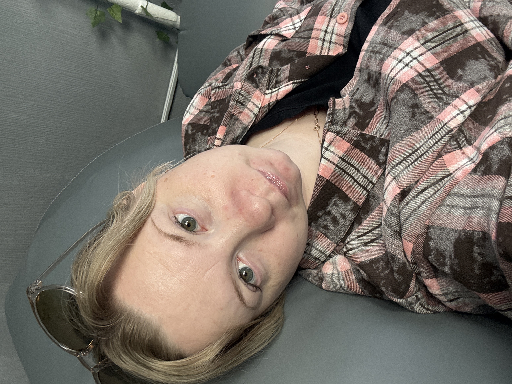 A person is lying down, wearing a pink and grey plaid shirt over a black top, and a gold necklace. Sunglasses are near their head. The location is not discernible as being in Chaikovsky, Russia, as there are no identifying landmarks or signage visible in the image. The environment consists of grey walls and a grey surface.