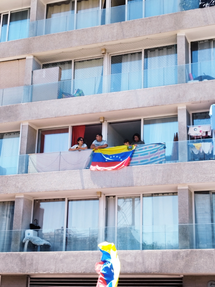 A multi-story building shows residents on a balcony, displaying a Venezuelan flag. Four people are visible, with the flag draped over the railing. Other balconies above and below feature glass railings and closed curtains. A smaller Venezuelan flag is seen in the foreground. The building's exterior is a light, textured gray, and the overall image is well-lit. Clothes hang from the adjacent balcony on the right.