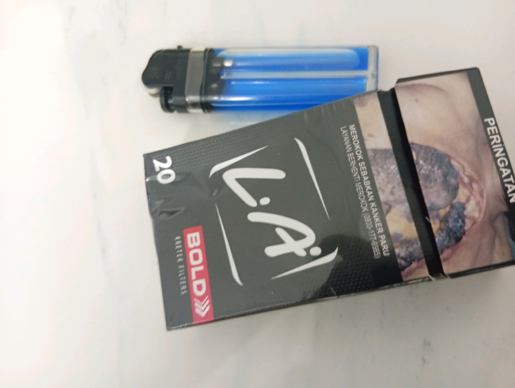 A pack of LA Bold Kretek Filters cigarettes and a blue disposable lighter lie on a white, textured surface, likely a countertop or table. The cigarette pack is predominantly black with a white stylized logo and "20" indicating the quantity. The lighter is transparent blue with black accents. The image appears to be an indoor setting, possibly a home or a shop in Samarinda, Indonesia. There are no people present in the image.