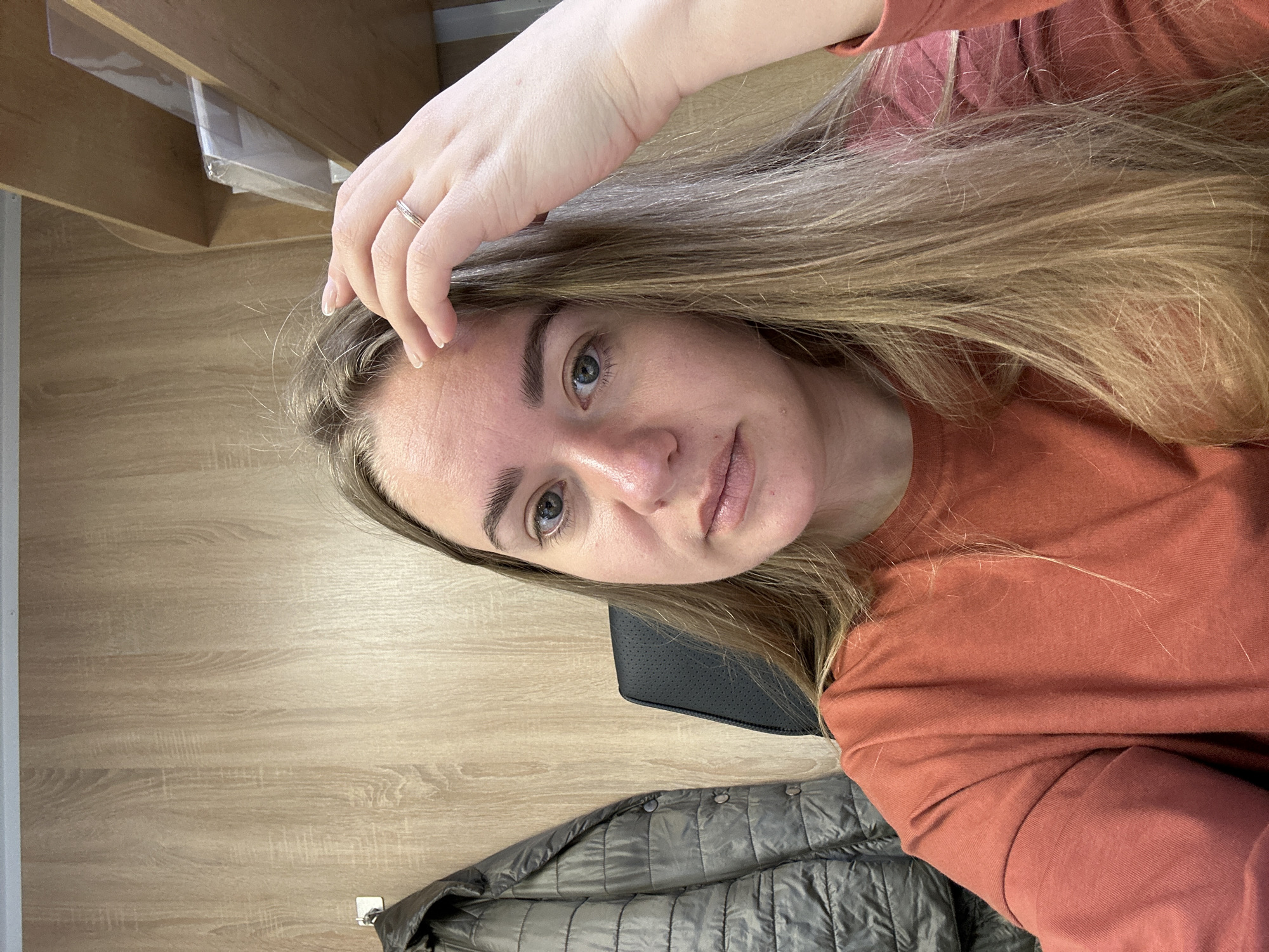 A woman with blonde hair and light complexion is visible in a selfie-style image. She is wearing a terracotta-colored long-sleeved shirt. Her right hand is raised, fingers touching her forehead. A silver ring is visible on her right ring finger.
The background includes a light brown wood-grain surface, a black chair, and a gray quilted jacket. Above the woman is a partially visible shelf.
There is no verifiable evidence of the specific location, Ural'skoe, Russia.