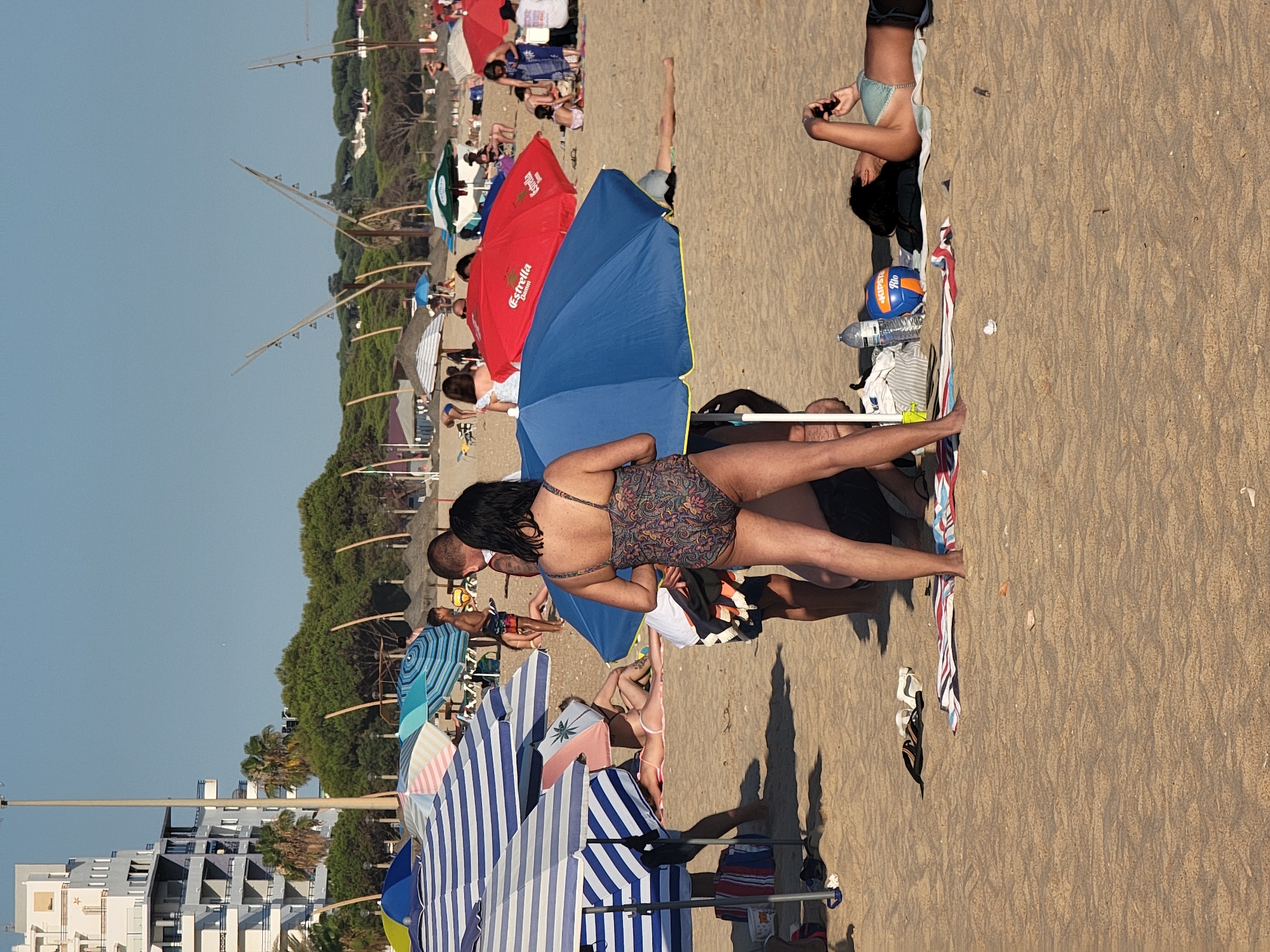 The image depicts a crowded beach scene in Quarteira, Portugal, under a clear blue sky. The foreground is dominated by sand, with several beach umbrellas providing shade to groups of people. In the center, a woman wearing a patterned one-piece swimsuit stands facing away from the camera, partially obscured by a large blue umbrella. To her right, another woman in a striped bikini lies on a towel, looking at her phone. Several other individuals are visible scattered across the beach, some sitting under umbrellas and others lying on towels. Beach accessories like towels, a volleyball, and a water bottle are present on the sand. In the background, a tall white building with multiple balconies stands out against a backdrop of green trees and vegetation. The overall scene captures a typical day at a busy summer beach.