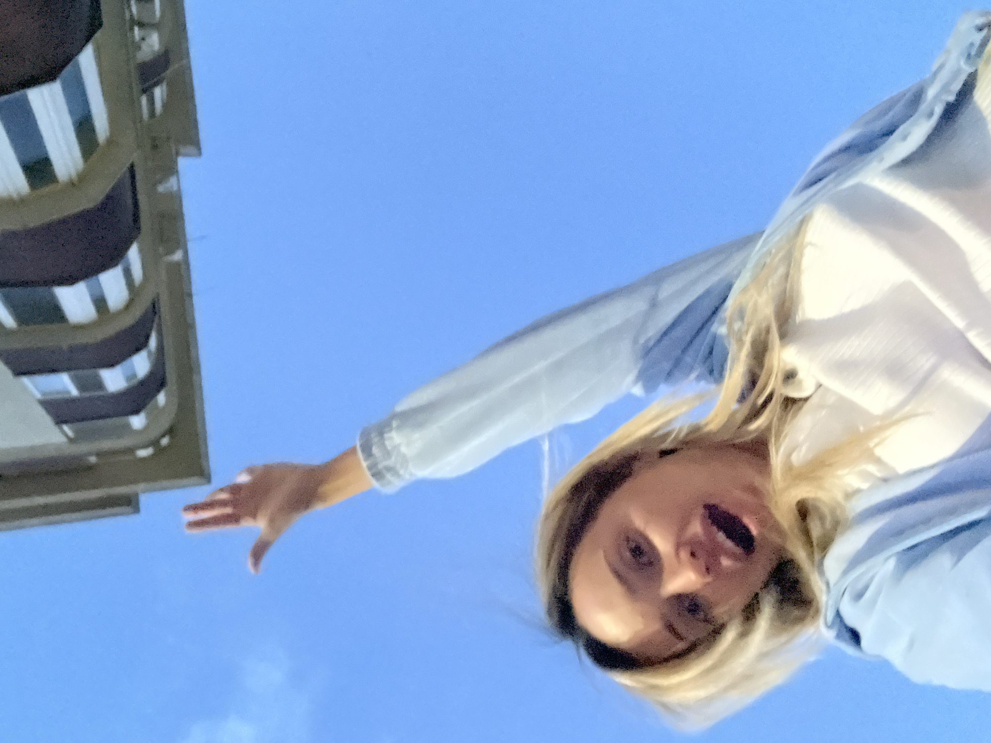 A young woman with blonde hair, wearing a light blue jacket over a white shirt, is pictured from a low angle, looking upwards towards the camera with her mouth open in an excited expression. Her right arm is raised, with her hand open and fingers spread, reaching towards a building structure that extends into the frame from the top. The background is a clear blue sky. The scene appears to be in Kryzhanivka, Ukraine, based on the metadata.
