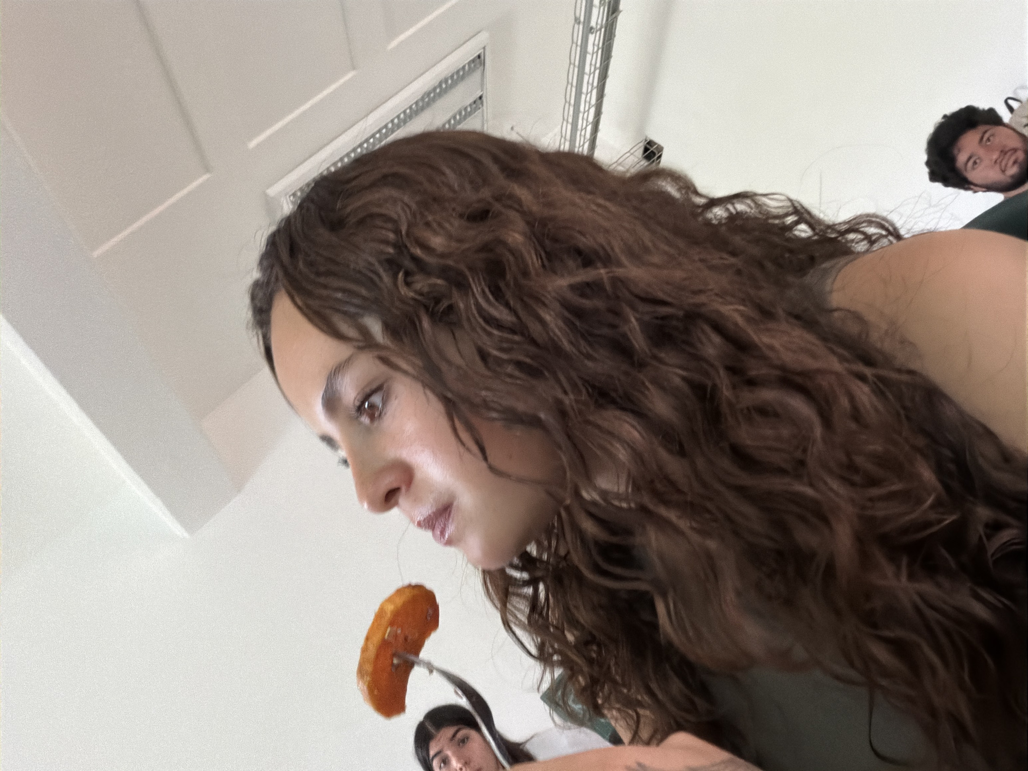 The image presents a low-angle, candid shot of three individuals in an indoor setting, likely in San Andrés Cholula, Mexico. The central figure is a young woman with long, wavy brown hair, her face in profile as she looks downwards. She holds a fork with a piece of orange-hued food, possibly a sweet potato or a plantain, poised near her mouth, indicating she is about to eat or has just taken a bite. Her expression is neutral and focused on the food.
In the lower mid-ground, partially obscured by the main woman's hand and the fork, another person with dark hair is visible, also looking upwards. Further in the background, towards the upper right corner, the face of a man with dark, curly hair and a beard is seen, looking towards the main woman with an observant or slightly surprised expression.
The background primarily consists of a bright white ceiling with architectural details, including square panels and a visible metallic vent or grating structure. The lighting is bright and even, suggesting it is daytime. The overall scene conveys a casual and informal gathering or mealtime moment. There is no visible text in the image.