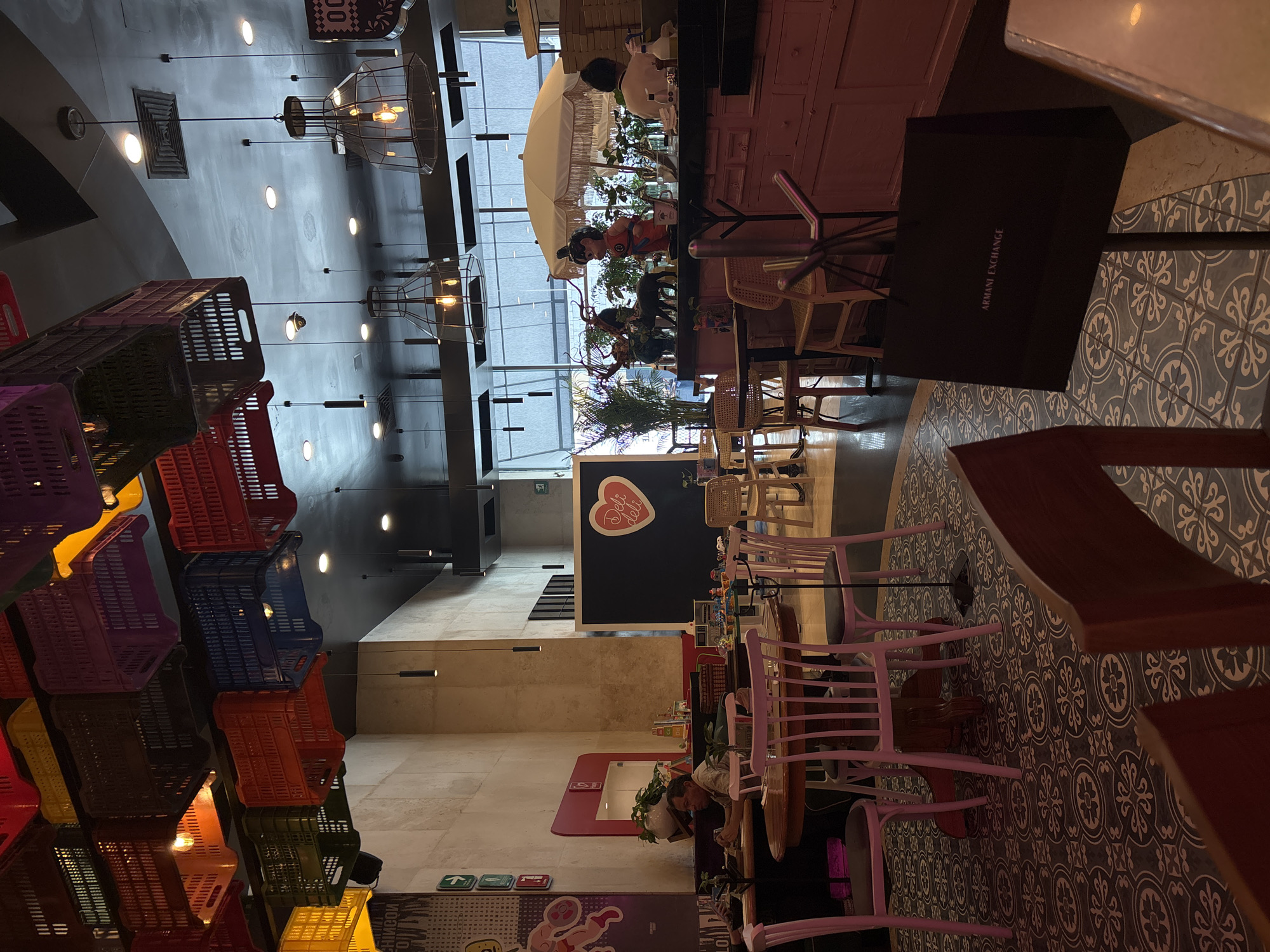 The image captures the interior of a restaurant in Puebla, Mexico, possibly a deli or cafe. The restaurant's decor is distinctive, featuring stacks of brightly colored plastic crates near the entrance, a high ceiling with modern hanging lights, and patterned tile flooring.
In the background, people are seated at tables near a window, shaded by a large white umbrella. A black Armani Exchange shopping bag is on a table in the foreground. Other tables with pink chairs are visible, and the "Deli Deli" logo is painted on a wall. The overall ambiance appears relaxed, possibly a mid-day scene during lunchtime.