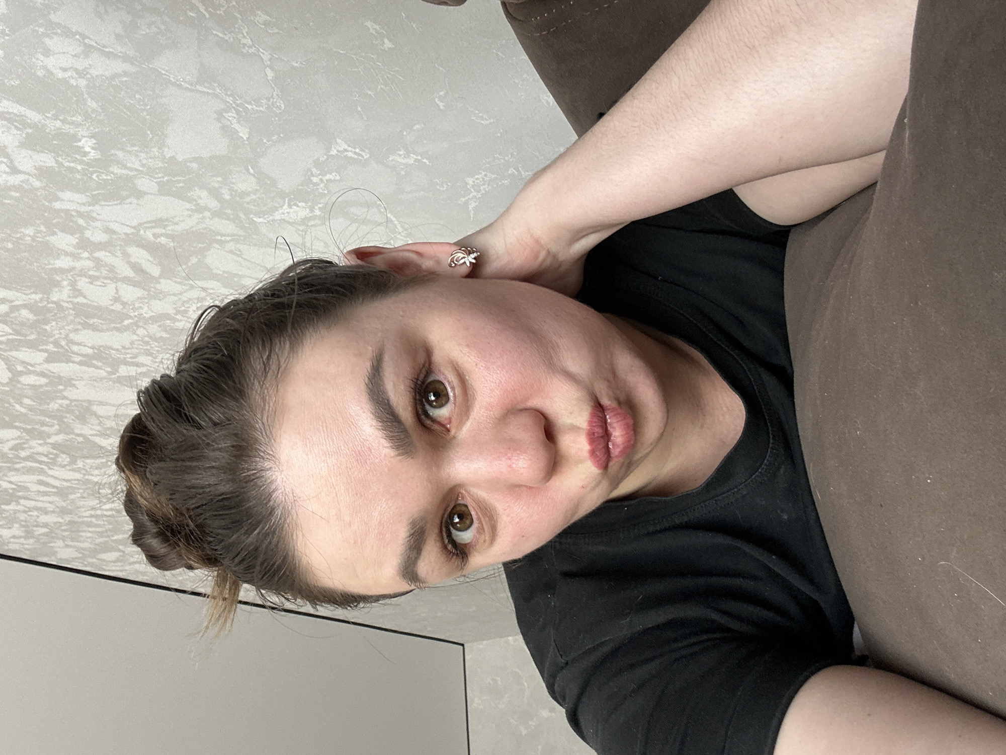 A woman is lying on a brown couch, wearing a black T-shirt, with her hair in a bun. She is holding her head with her right hand, wearing a sparkly earring in her right ear. The background consists of a textured grey wall and a white door frame. This image does not allow for the determination of a specific location in the city of Chaikovsky, Russia.