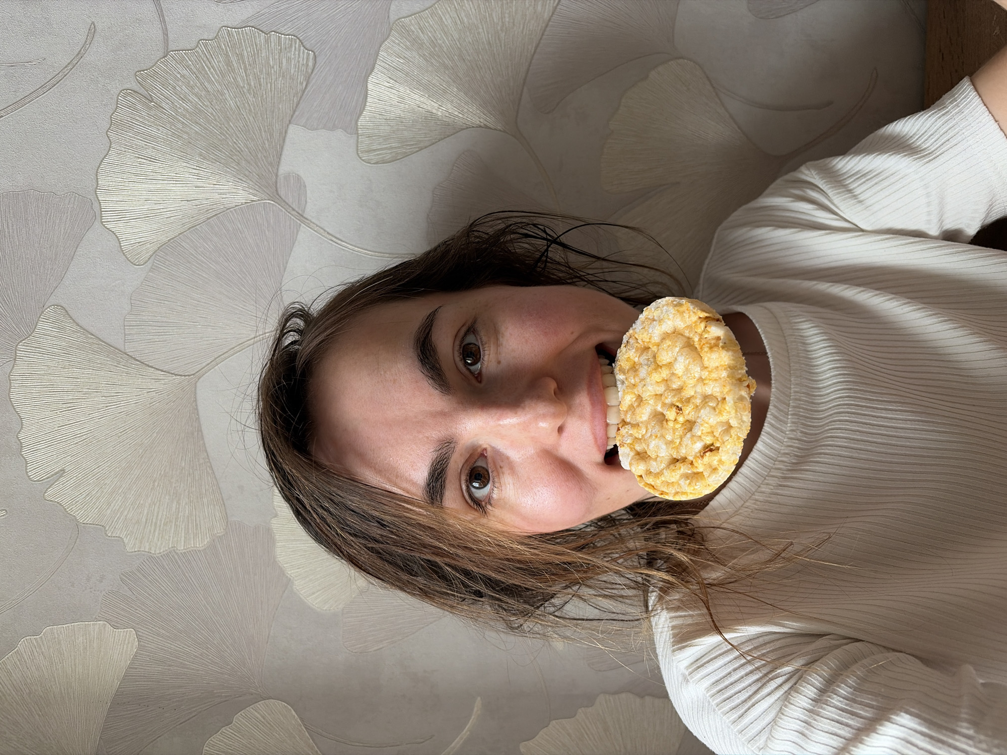A woman is holding a rice cake in her mouth. The woman has brown hair and brown eyes and is wearing a white, ribbed top. The background is a wall with a repeating pattern of stylized ginkgo leaves in shades of gray. The image is rotated, with the woman's head oriented towards the left side of the frame. The location is not discernable from the image.