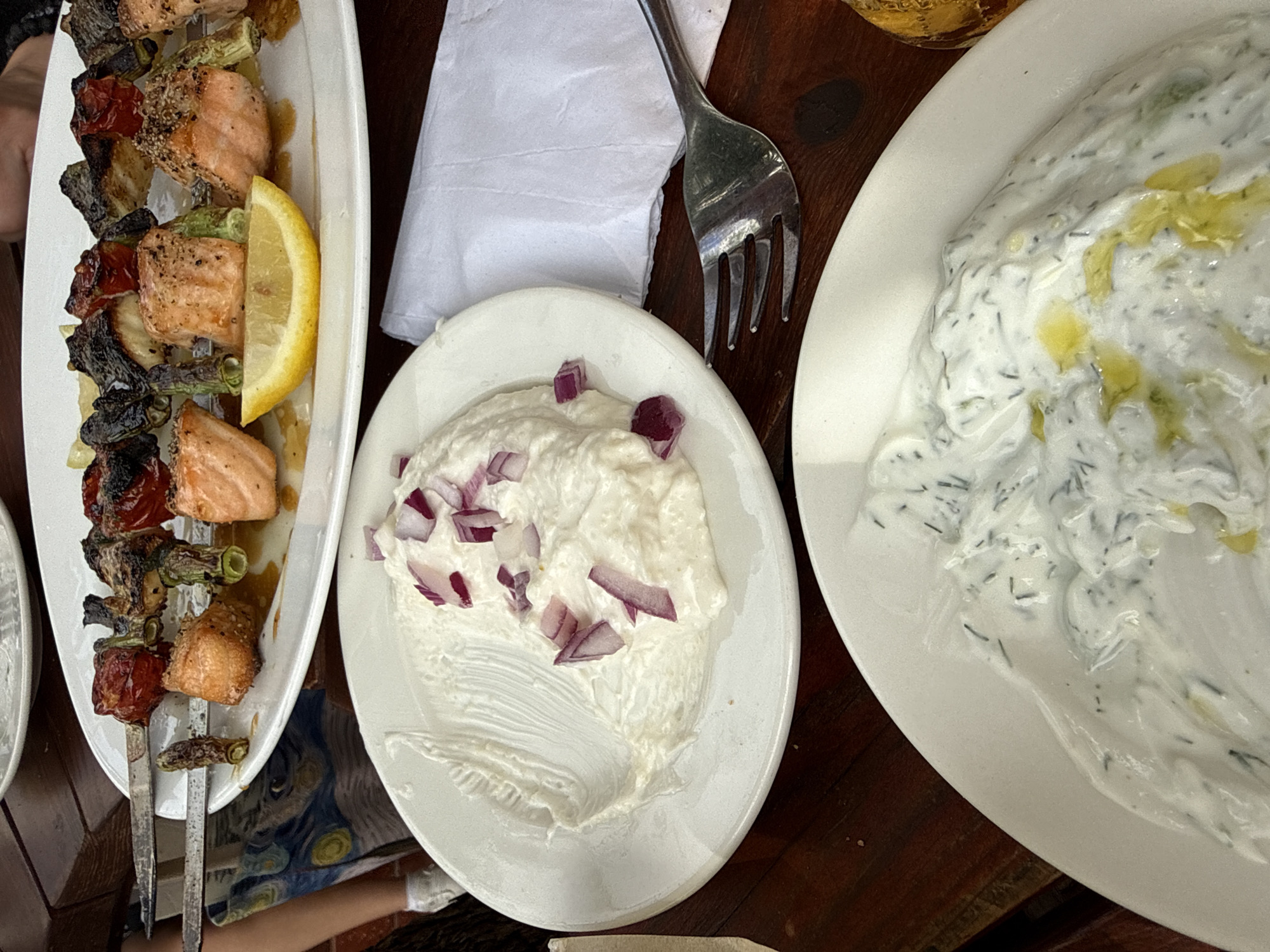 The image presents a top-down view of a dining table arrangement. Dominating the scene are three white plates: one contains salmon skewers with grilled vegetables and a lemon wedge; another plate holds a white creamy dip topped with chopped red onions; and the third plate features a white creamy dip with dill. A silver fork rests near the center plate, partially on a white napkin. The table appears to be made of wood. The scene appears to be in Tel-Aviv, Israel, based on the menu and style of food served.
