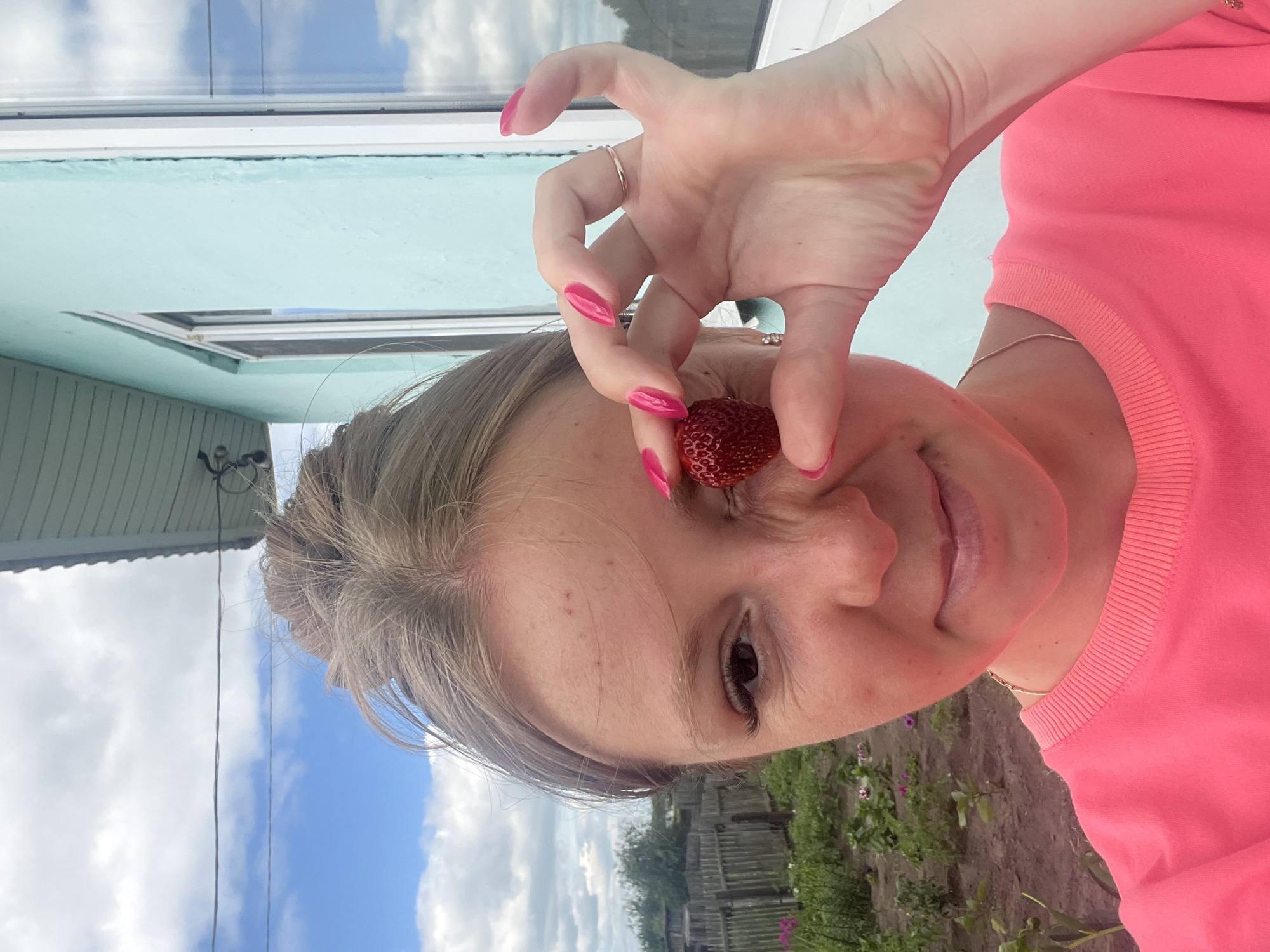 A young Caucasian woman with blonde hair tied in a messy bun is taking a selfie outdoors. She is wearing a bright pink sweatshirt. Her right eye is partially obscured by a ripe red strawberry, which she is holding with her right hand. Her fingernails are painted a vibrant pink. In the background, there is a light blue building with a white-framed window, a wooden fence, and a garden with green plants and some purple flowers. The sky is cloudy with patches of blue. The scene appears to be in a rural or suburban area, possibly in Сухринское, Russia, given the architectural style and vegetation.