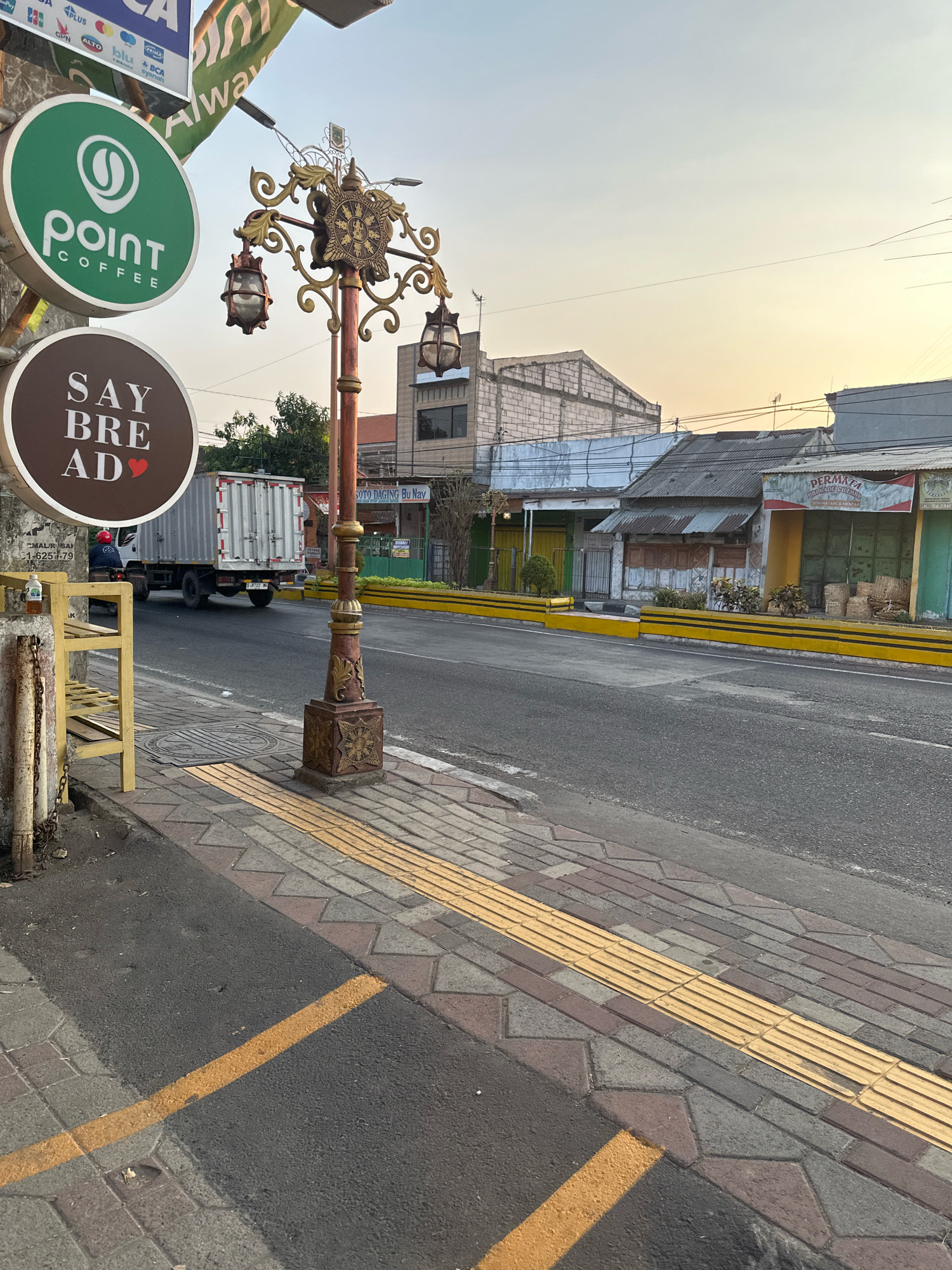 The image depicts a street scene in Mojokerto, Indonesia. On the left, two circular signs are visible: a green one with a white "point coffee" logo and a brown one with white text "SAY BRE AD" and a red heart. A decorative streetlamp with three lanterns stands in the foreground. A white truck is driving on the road. The sidewalk has tactile paving for the visually impaired. The background shows buildings and shops, some with faded signs, and a yellow barrier separating the sidewalk from the road. The sky is clear with a warm sunset glow.