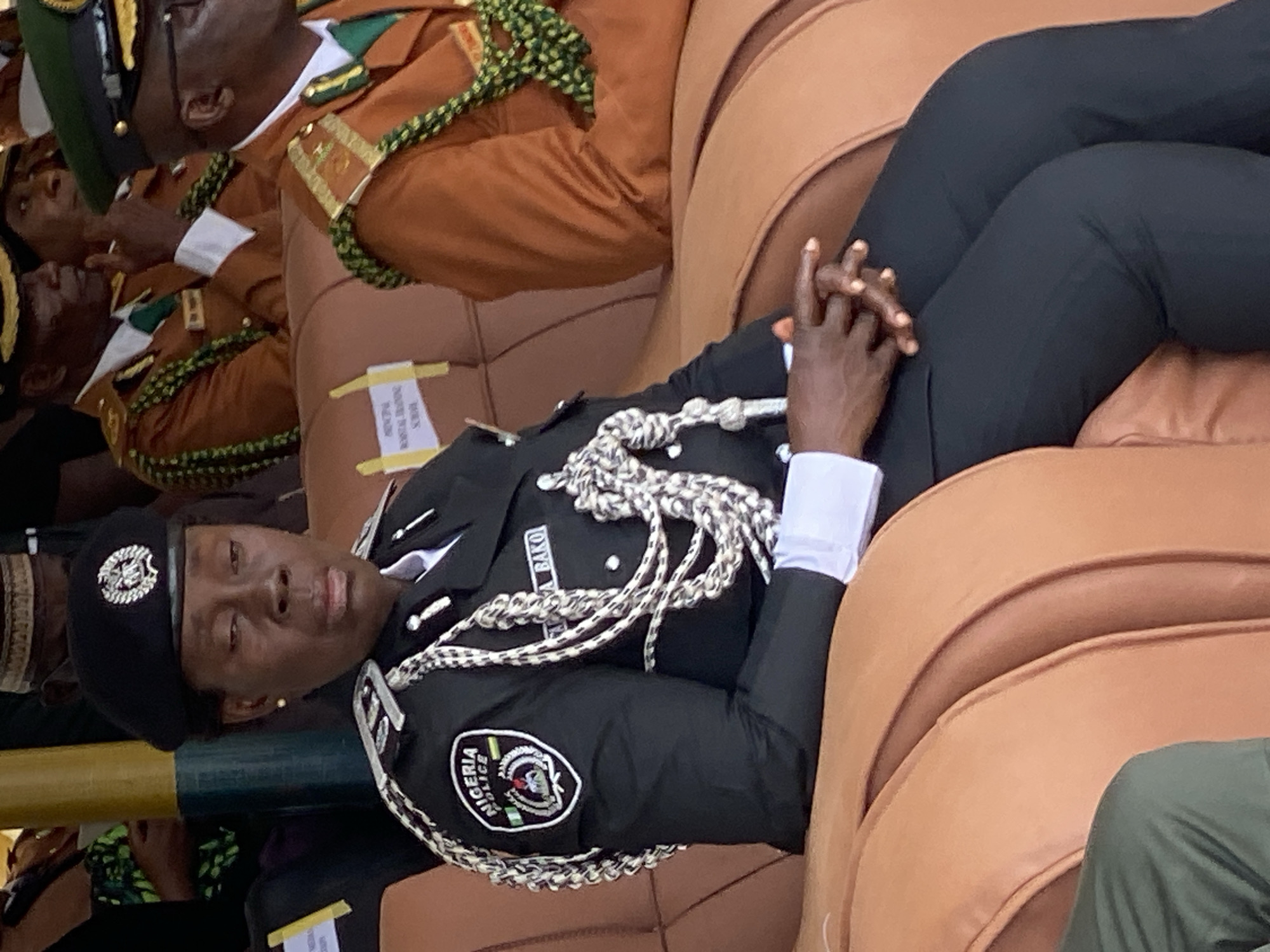 The image shows a female police officer in uniform, sitting in a brown upholstered seat. She is wearing a black beret with a police emblem, a black jacket with silver braiding and a Nigerian Police Force patch on the left sleeve, and black trousers. She has her hands clasped in her lap. In the background, other individuals in uniform are visible, some wearing brown jackets with gold trim. The setting appears to be an official event, likely in a formal seating area. The name "BAKO" is visible on her jacket. The event is taking place in Kaduna, Nigeria.