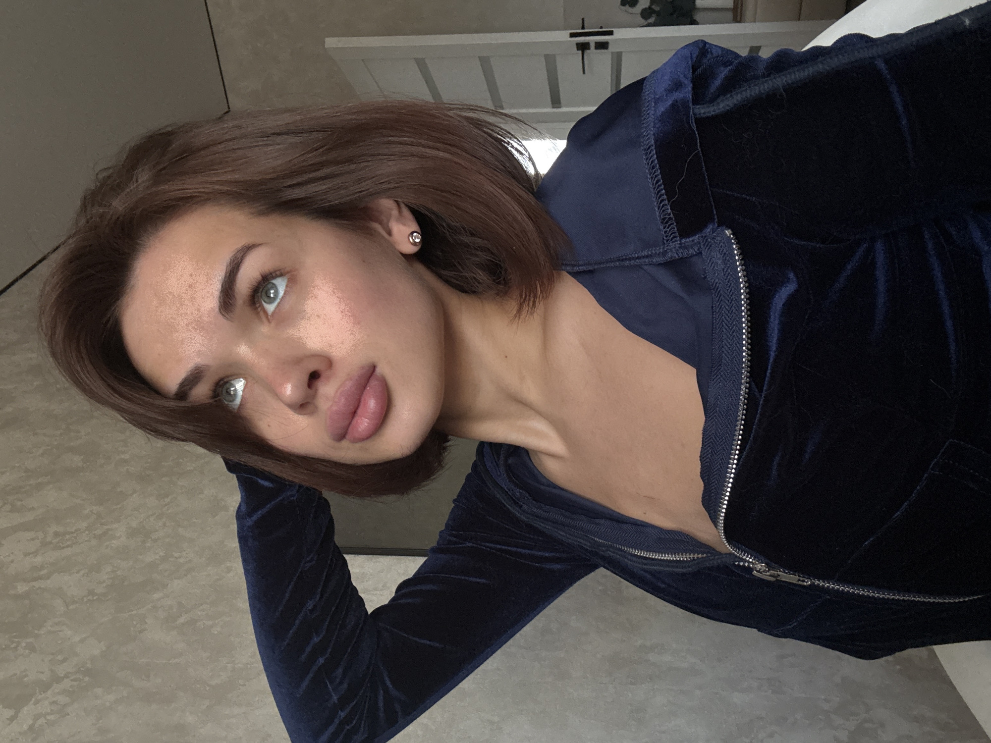 The image shows a woman indoors. She has light skin, brown hair, and light-colored eyes. She is wearing a dark blue velvet top with a zipper. The background includes a wall and part of a white structure, possibly a door or furniture. The woman is positioned in a way that suggests she may be taking a selfie. There is no explicit information indicating the image was taken in Лобаново, Russia.