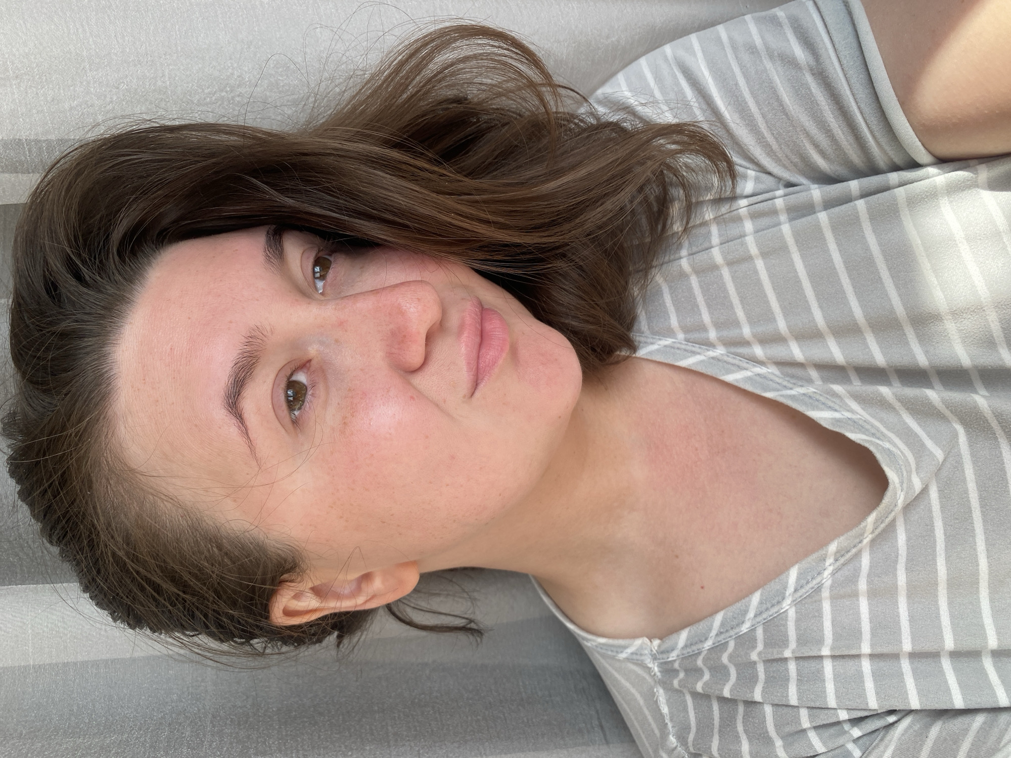 The image features a close-up portrait of a woman, appearing to be reclining or lying down, with the image rotated 90 degrees clockwise. She has medium brown, wavy hair that frames her face and flows loosely. Her eyes are brown, and she looks directly towards the viewer with a neutral, relaxed, and slightly contemplative expression. Her lips are a natural pink color, slightly parted.
Her complexion is fair, with noticeable freckles across her nose and cheeks. There is some subtle redness visible on her neck and upper chest. She is wearing a casual top with light grey and white vertical stripes. The background consists of a light-colored, textured fabric, possibly a curtain or bedding, suggesting an indoor, domestic setting in Chaikovsky, Russia.
The lighting appears soft and natural, indicating daylight. There is no visible text in the image. The crops provide closer views of her eyes, lips, the texture of her skin and hair, and the striped pattern of her clothing, confirming these details and highlighting the subtle skin redness on her neck and chest area.