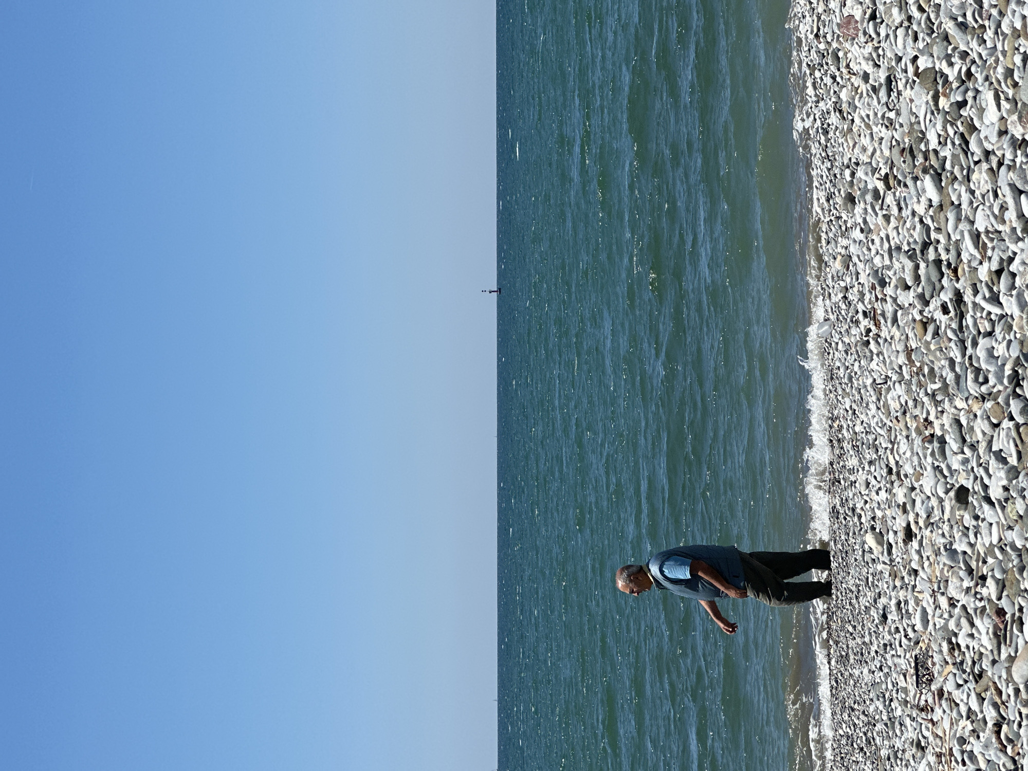 A vertically-oriented photograph shows a man on a pebble beach next to the ocean. The sky is clear blue. The ocean is dark blue with visible ripples. The man is wearing a blue shirt and dark pants. He is walking along the edge of the water. The beach is covered in grey and white pebbles. In the distance, a small dark vertical shape, likely a buoy or marker, sits on the water's surface. The location is likely the city of Fethiye, Turkey, based on the prompt.