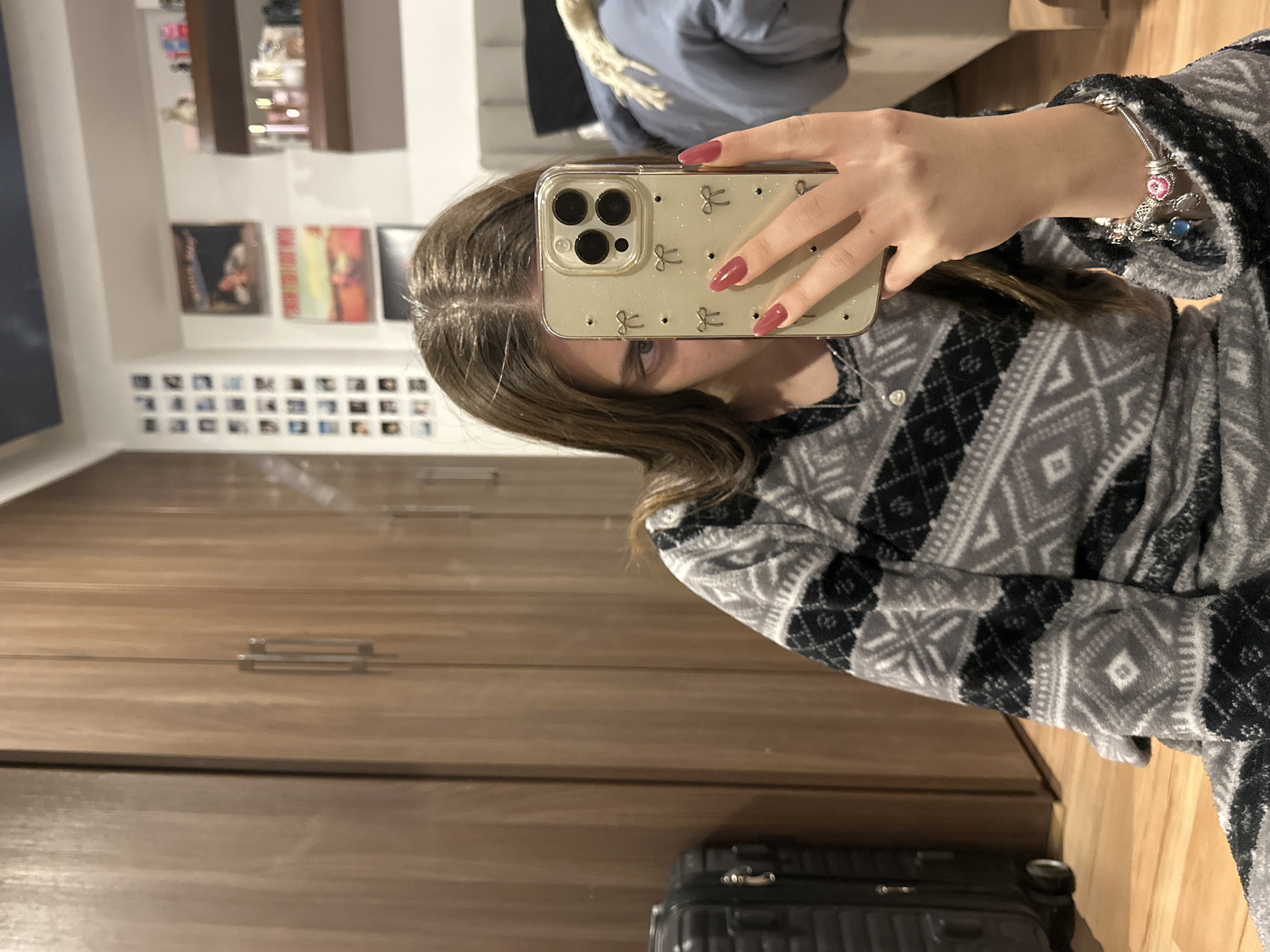 A person is taking a selfie with an iPhone in a bedroom. The person is wearing a gray and black patterned pajama top and has reddish-pink nail polish. A silver charm bracelet is visible on their wrist. The background includes a wooden wardrobe, artwork, and photographs on the wall. A suitcase is visible on the floor. The location is likely a residential area, potentially within the city of Huixquilucan de Degollado, Mexico, based on the general setting.