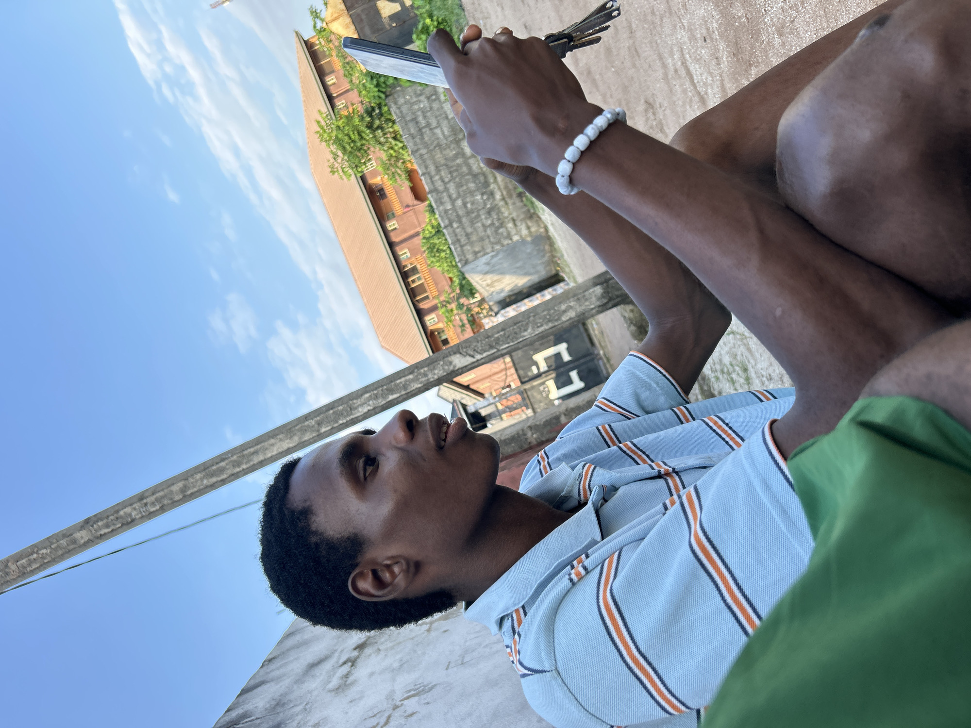 A young Black male is sitting outdoors, holding a smartphone in his right hand and a set of keys in his left. He is wearing a light blue collared shirt with thin orange and dark blue stripes. A white beaded bracelet adorns his left wrist. He appears to be looking up and to his left, with a relaxed expression.
The setting suggests an urban or semi-urban environment in Ihiagwa, Nigeria. In the background, a multi-story building with reddish-brown siding and white trim is visible, partially obscured by lush green trees. The sky is clear blue with scattered white clouds, indicating fair weather, likely during the daytime. The foreground shows a concrete surface where the man is seated, with a portion of green fabric on the right side of the frame, possibly clothing. The image is taken from a low angle, looking up at the subject and the sky. There is no visible text in the image.
