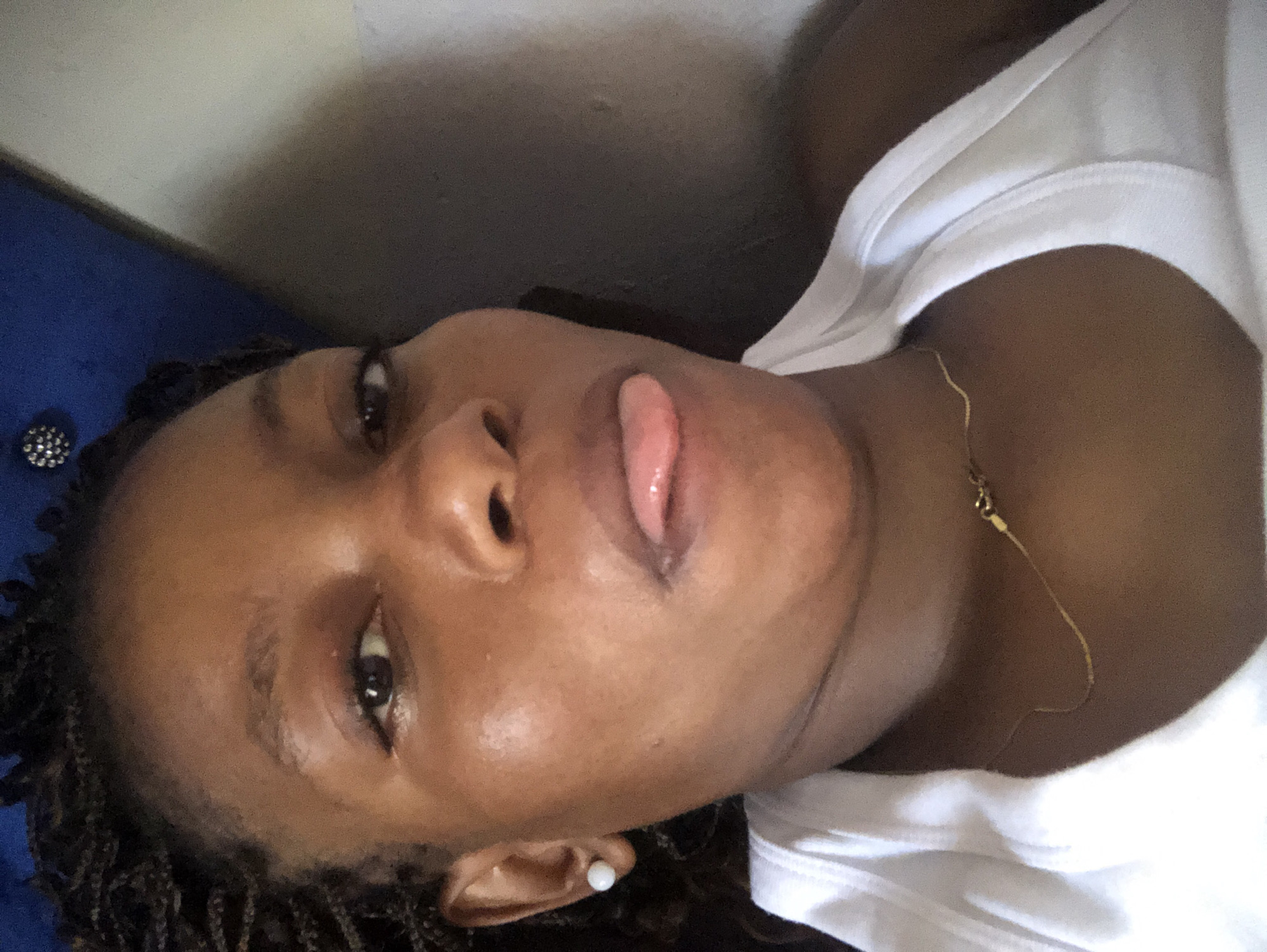 A close-up, slightly angled view depicts a Black woman with dark brown skin and small braids laying on a blue upholstered surface. She is wearing a white t-shirt and a gold necklace. Her lips are slightly parted, revealing a glimpse of her tongue. She has a white stud earring in her right ear. A decorative button is visible on the blue upholstered surface behind her. The location is Kashere, Nigeria.