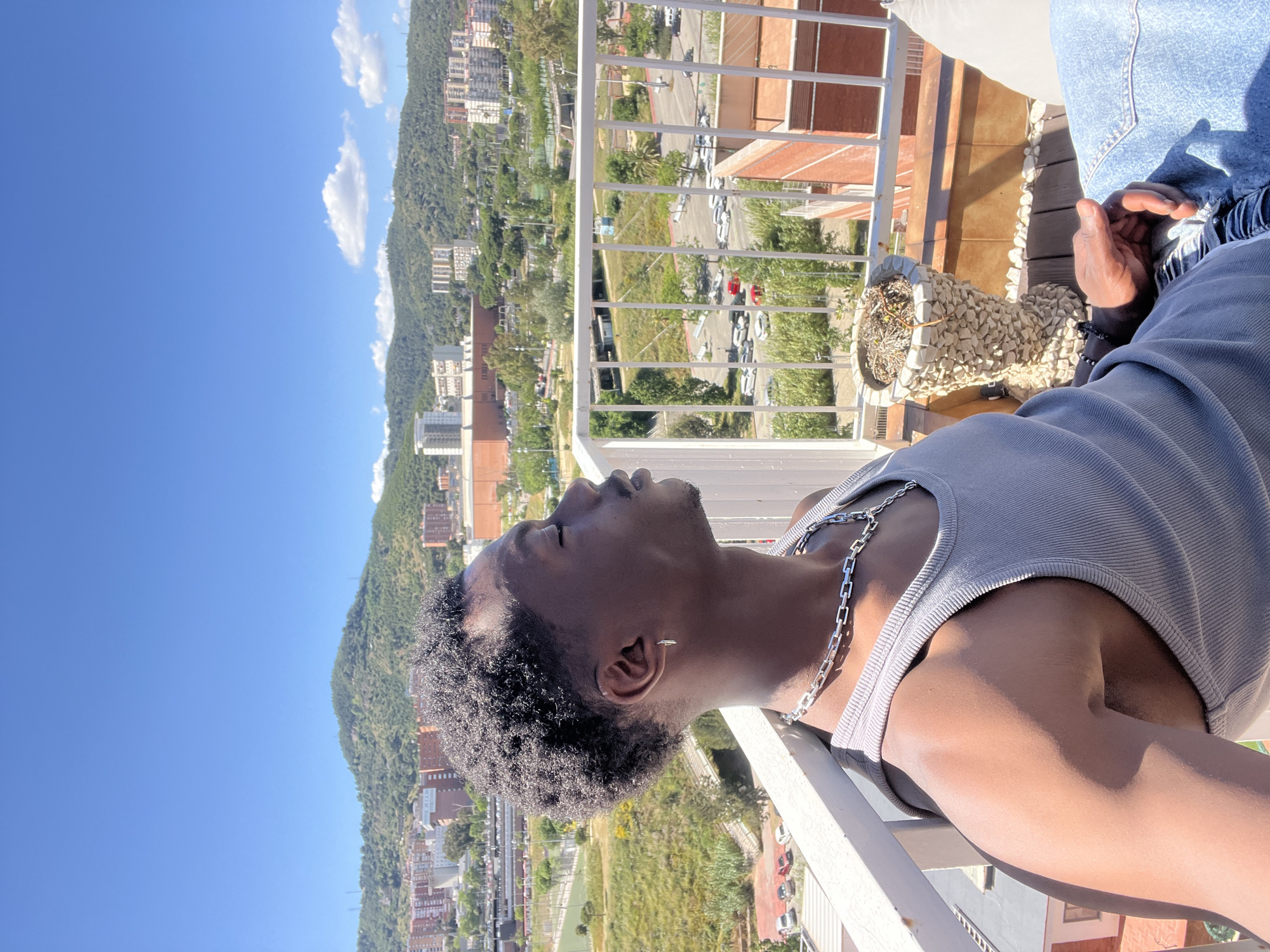 A dark-skinned person, likely male, is lying on a white-painted balcony railing with their head tilted back and eyes closed. He wears a grey tank top and a silver chain necklace. The balcony overlooks an urban landscape featuring green spaces, buildings, and a mountain ridge in the background under a bright blue sky. The image does not depict Barcelona, Spain, based on the architecture and geographical features. The specific location is not identifiable.