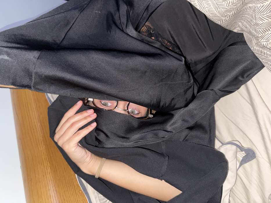 A woman lies in bed under a black garment, eyes visible through glasses. The dark material is draped over her head, partially obscuring her face. One hand, adorned with a simple wristband, is visible at the bottom of the frame. A hint of lace is visible in the upper part of the black fabric. The bedsheets are patterned, and the headboard of the bed is partially visible on the left side. The photo's orientation appears to be flipped.