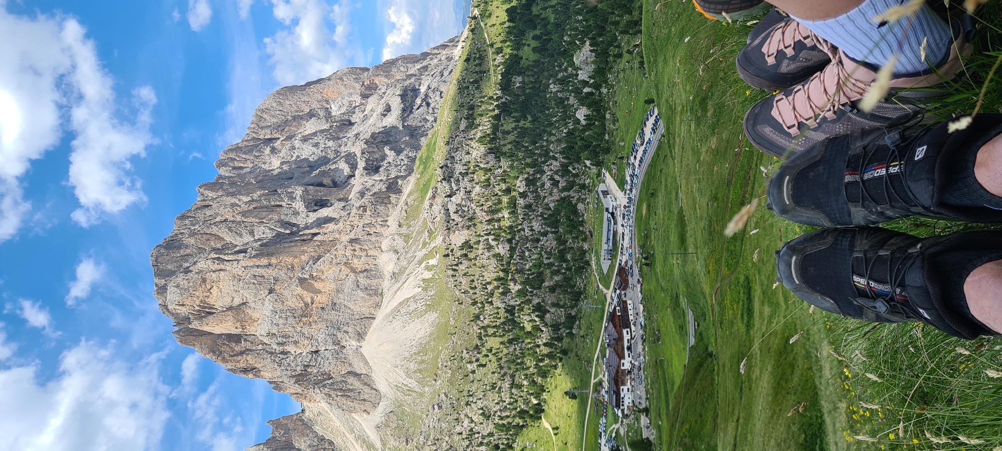 The image shows a scenic view of the Dolomites in Val Gardena, Italy, with a prominent, rugged mountain peak dominating the background. The mountain is largely devoid of vegetation, displaying its rocky, eroded face under a bright blue sky with scattered white clouds. In the mid-ground, a green, rolling landscape is covered with sparse trees and bushes, leading down to a developed area with buildings, including a large lodge or hotel, and extensive parking lots filled with vehicles. A road curves through the area. The foreground features the lower legs and feet of two individuals sitting on a grassy slope, with their hiking boots visible. One pair of boots is black with red and yellow accents, and the other is a lighter color with pink laces. Blades of grass and some small yellow flowers are present in the immediate foreground. The overall scene suggests a recreational outdoor setting, likely a hiking or tourist destination.