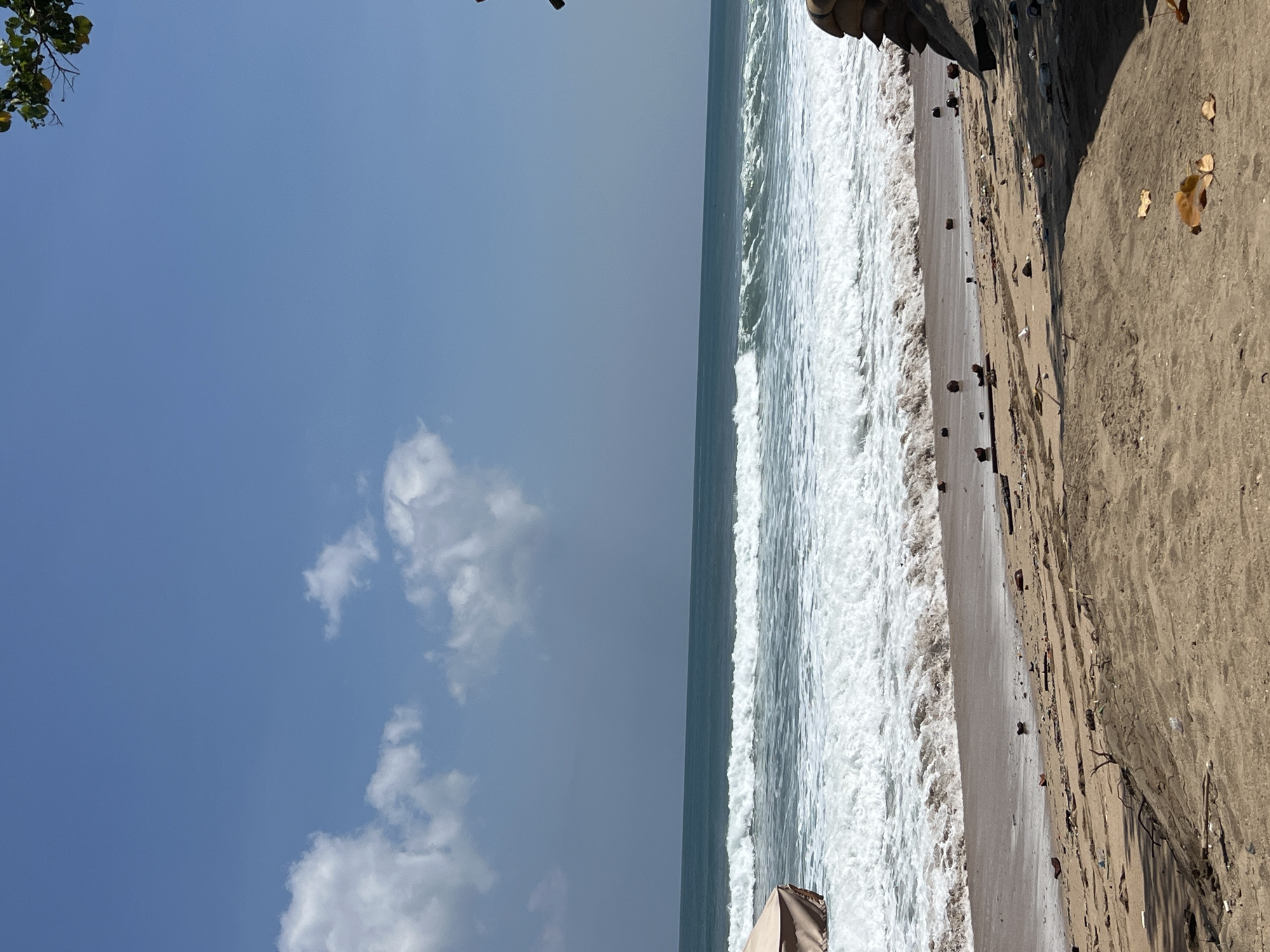 An outdoor, eye-level, full shot captures a sandy beach in Kuta, Indonesia, under a clear, bright blue sky with scattered white clouds. The ocean is a deep teal with prominent white waves rolling towards the shore. The beach itself is littered with debris, including coconuts, sticks, and what appears to be plastic waste. A portion of a beige beach umbrella is visible on the left side of the frame, and a pile of sandbags is stacked on the right. Sunlight casts shadows across the sand, indicating a bright, sunny day.