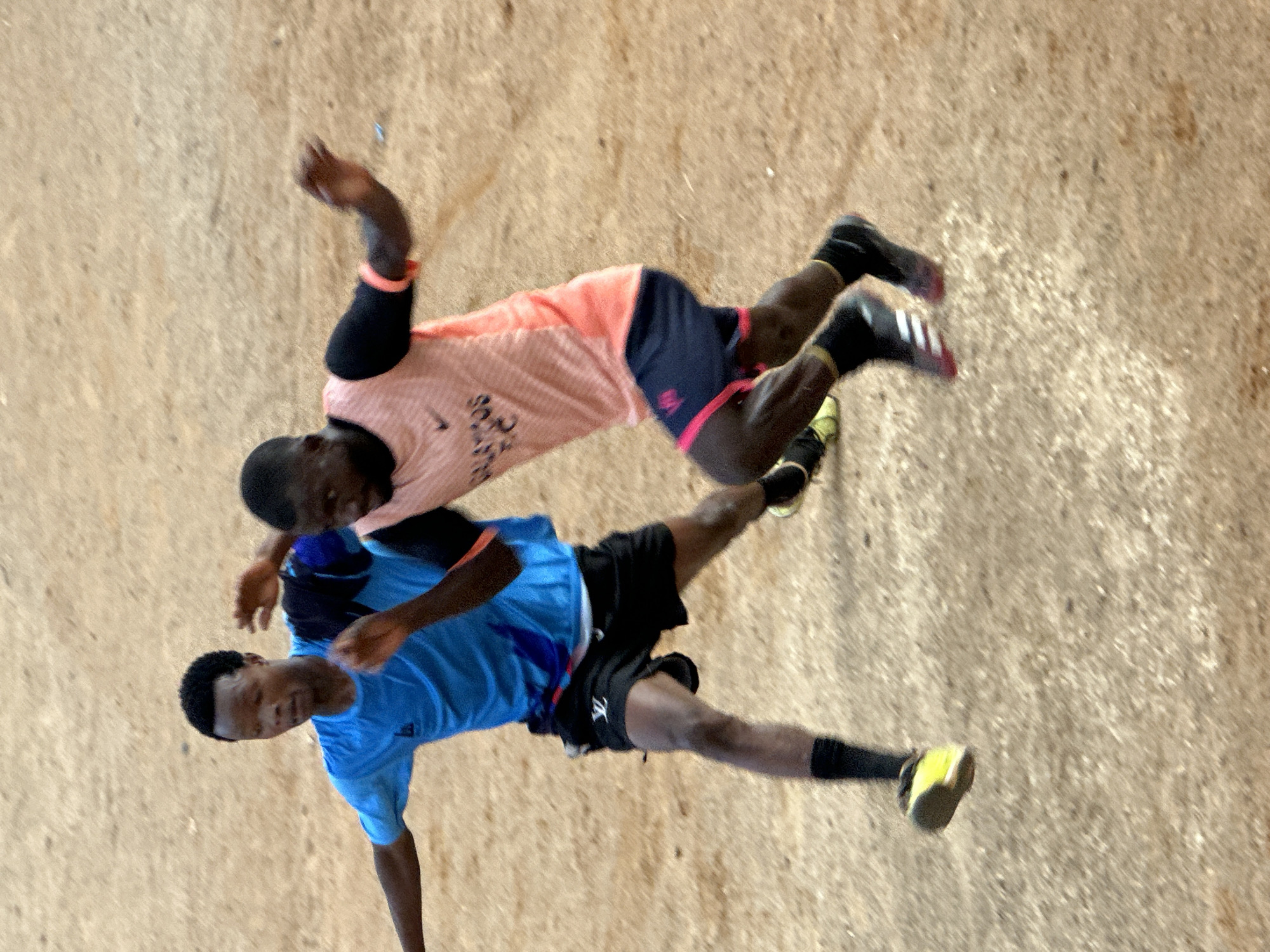 Two young Black men in athletic wear are engaged in a dynamic activity on a sandy or dirt surface, possibly a sports field. The man on top is wearing a pink and orange t-shirt with black trim and dark blue shorts. He has black arm sleeves and is in motion, with his legs extended. The man below is wearing a bright blue t-shirt and black shorts with red trim. He is also in motion, with his body low to the ground and one leg extended, almost as if he's attempting to tackle or block the other player. Both men are wearing athletic shoes, with the lower man's shoes appearing to be neon yellow and black.
The image is taken from a slightly elevated, overhead perspective, and the lighting suggests it is daytime, with no indication of specific weather conditions. The ground appears dry and uneven, consistent with an outdoor playing area in a location like Jos, Nigeria. The intensity of their stances and the blur of motion indicate an energetic and competitive interaction, likely a sport like football (soccer). There is a sense of athleticism and focused effort displayed by both individuals. Some text is visible on the t-shirt of the man on top, but it is largely obscured and illegible. The overall impression is one of active play in a casual, outdoor environment.
