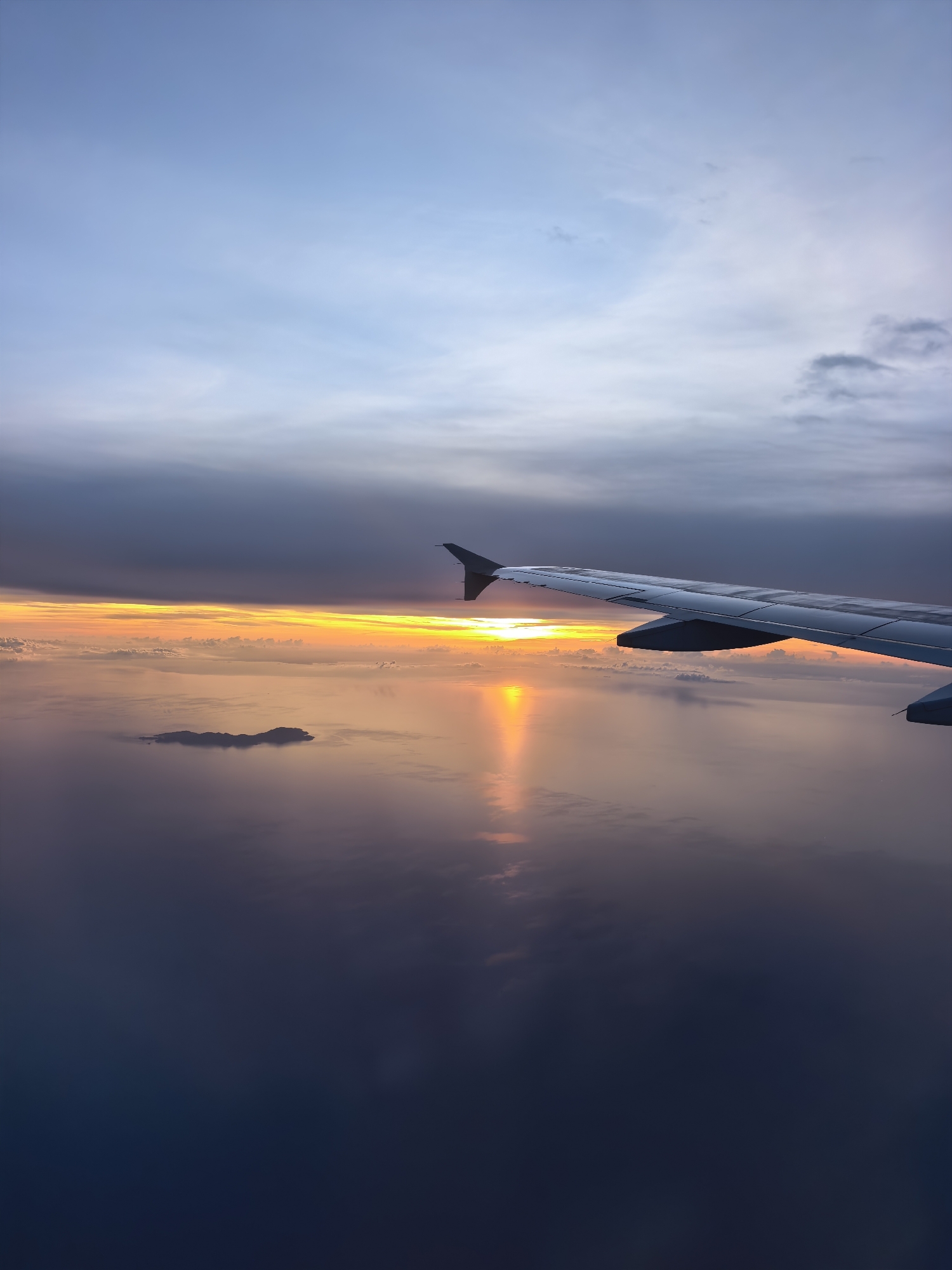 An aerial view captures a sunset over a vast body of water, with a small island visible in the distance. The sun's golden rays reflect on the water's surface, creating a shimmering path. The foreground is dominated by the wing of an airplane, angled upwards, suggesting the viewer is a passenger. The sky transitions from a soft blue at the top to darker, cloud-filled layers closer to the horizon, where the vibrant orange and yellow hues of the sunset are most intense. The scene appears serene and peaceful, bathed in the warm light of the setting sun.