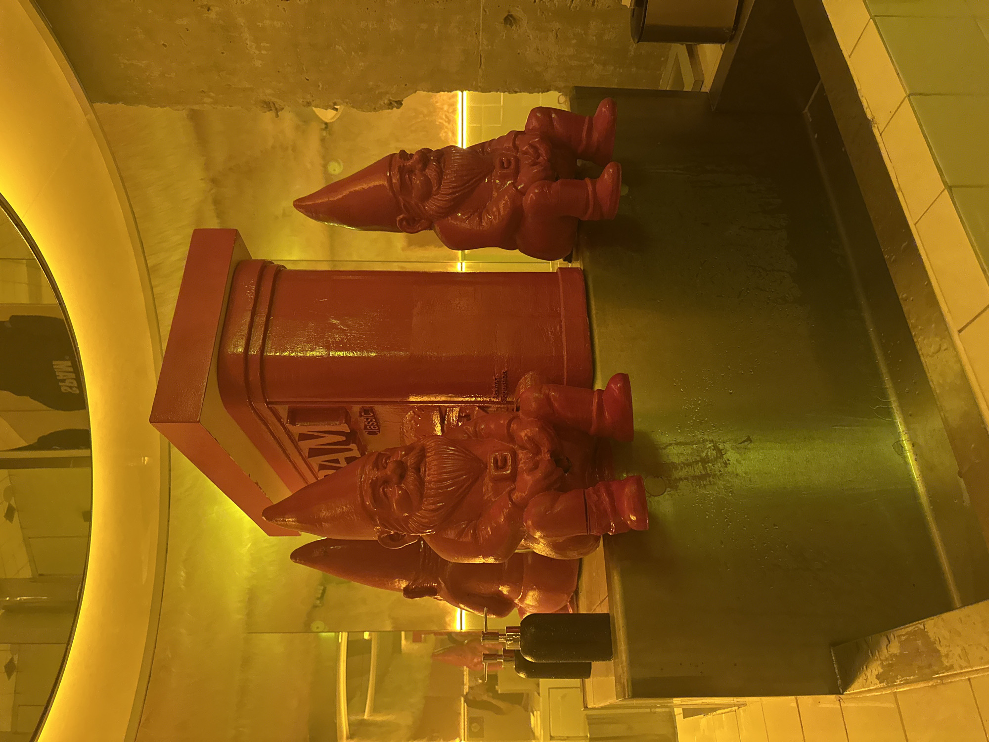 The image depicts a sink area in what appears to be a restroom, possibly in Mexico City, Mexico, as indicated by the SPAM-branded decoration. Two sets of red-painted gnome figures are placed around a red SPAM promotional item. The sink itself is metallic and reflective, and the lighting is yellow-toned. Two black soap dispensers are installed above the sink surface. The surrounding wall consists of concrete and tile. A mirror with a yellow frame is partially visible on the left side of the image.