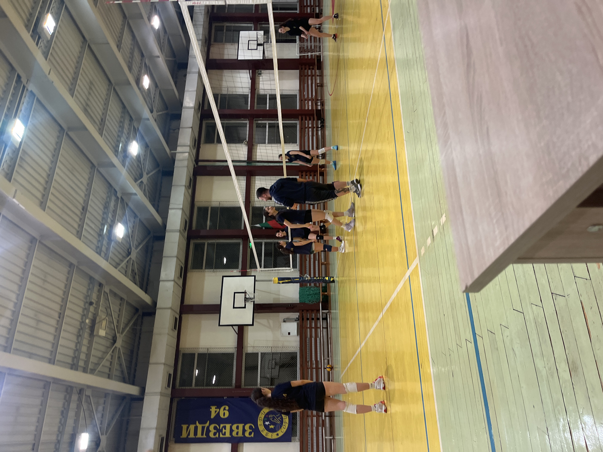 A volleyball team practices in an indoor court, likely located in Sofia, Bulgaria, as indicated by the "ЗВЕЗДИ 94" banner. Players, wearing dark jerseys, are positioned near the volleyball net, along with a coach. The court has yellow and green sections with line markings. Basketball hoops are visible in the background. The facility has windows along the sides and a metal ceiling structure with lights.