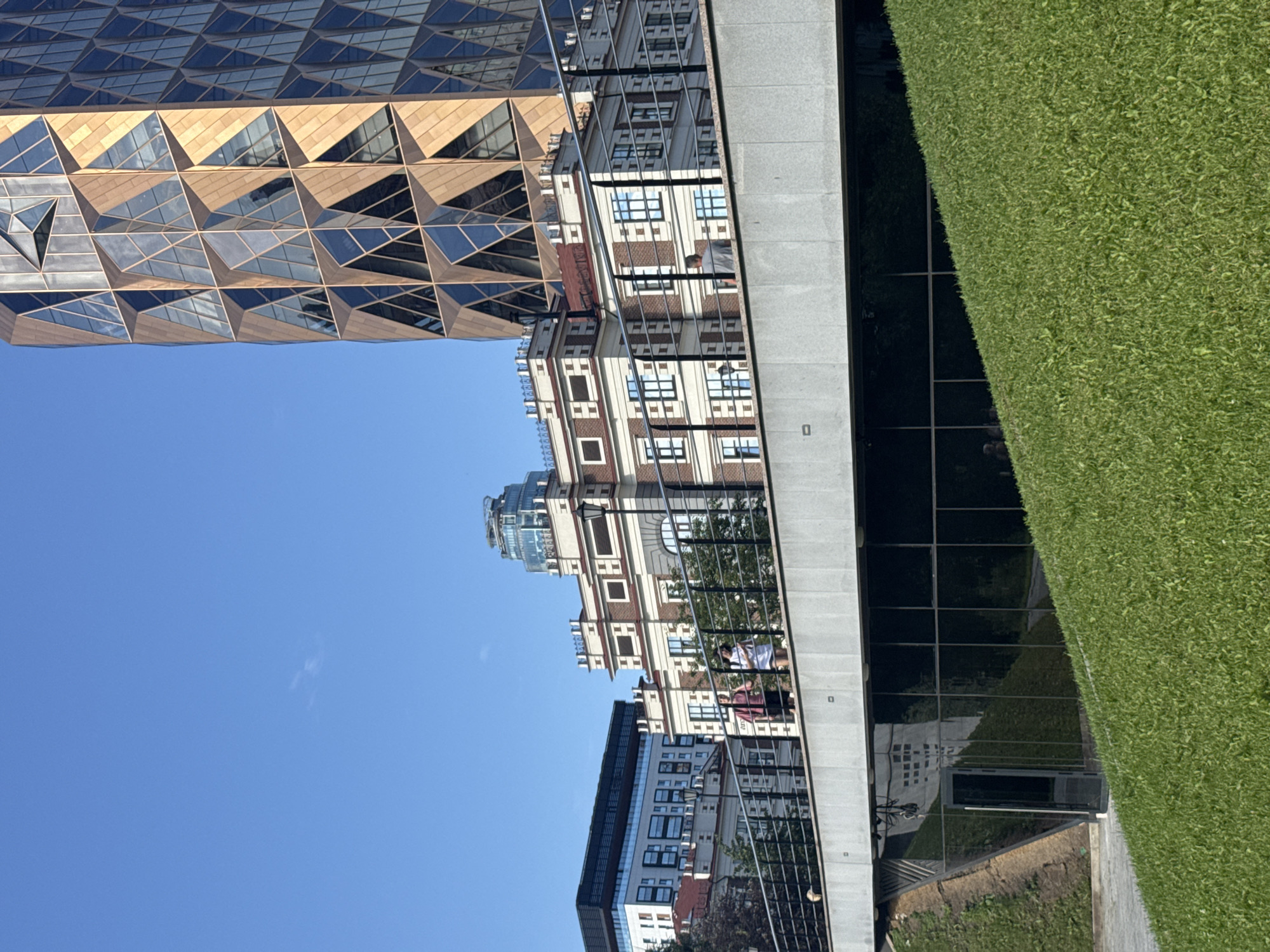 The image shows a sunny day in Yekaterinburg, Russia, with a clear blue sky. In the foreground, a sloped green lawn leads up to a modern building with a glass facade and a door. Above this, a pedestrian bridge spans across the frame, with a metal railing. Two people are visible walking on the bridge. Behind the bridge, a classical-style building with red and white brickwork and white trim stands prominently. Further in the background, a modern skyscraper with a distinctive geometric design and a golden facade rises into the sky. Another building with a dark, horizontal facade and many windows is visible to the left. A round-topped building with a glass dome is seen further in the distance. The overall scene depicts a mix of architectural styles within an urban setting.