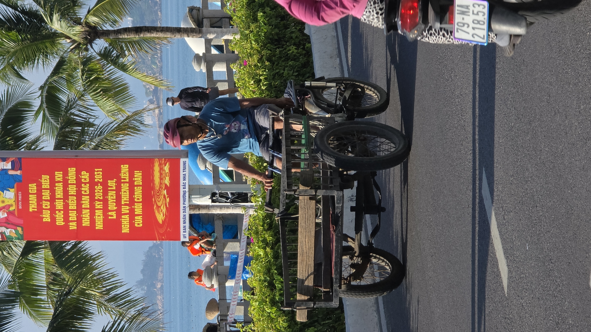 A sideways view captures a street scene in Nha Trang, Vietnam. A man is on a modified tricycle with a wooden platform attached, likely used for carrying goods. The tricycle is positioned on an asphalt road marked with a white line. Behind him, partially obscured, is a motorcycle. A license plate reading "79-NA 218.53" is visible on the motorcycle. A sign is displayed to the left, with Vietnamese text regarding participation in elections, and mention of "ỦY BAN NHÂN DÂN PHƯỜNG BẮC NHA TRANG" ("People's Committee of Bac Nha Trang Ward"). Behind the sign is a row of greenery, and in the distance, a body of water is visible, as well as palm trees and a partial view of a structure along the waterfront. The lighting suggests a sunny day. Several individuals in orange vests are sitting next to the barrier and the sign.