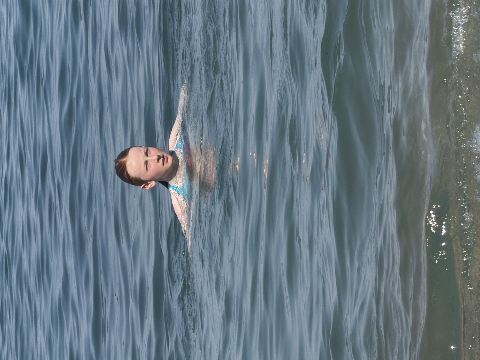 In a top-down perspective, a young person with fair skin and red hair is partially submerged in water. Their head is tilted back, eyes closed, and their mouth is open as if in mid-breath or exhalation. They are wearing a patterned swimsuit with blue and pink elements. The water is a calm, blue-grey, with distinct ripples and waves creating a textured surface. Sunlight glints off the water in the upper right portion of the image, suggesting a bright, possibly sunny day. The overall impression is one of peaceful immersion in the water. The provided crops offer closer views of the subject's face, hair, and swimsuit, as well as different sections of the rippling water.