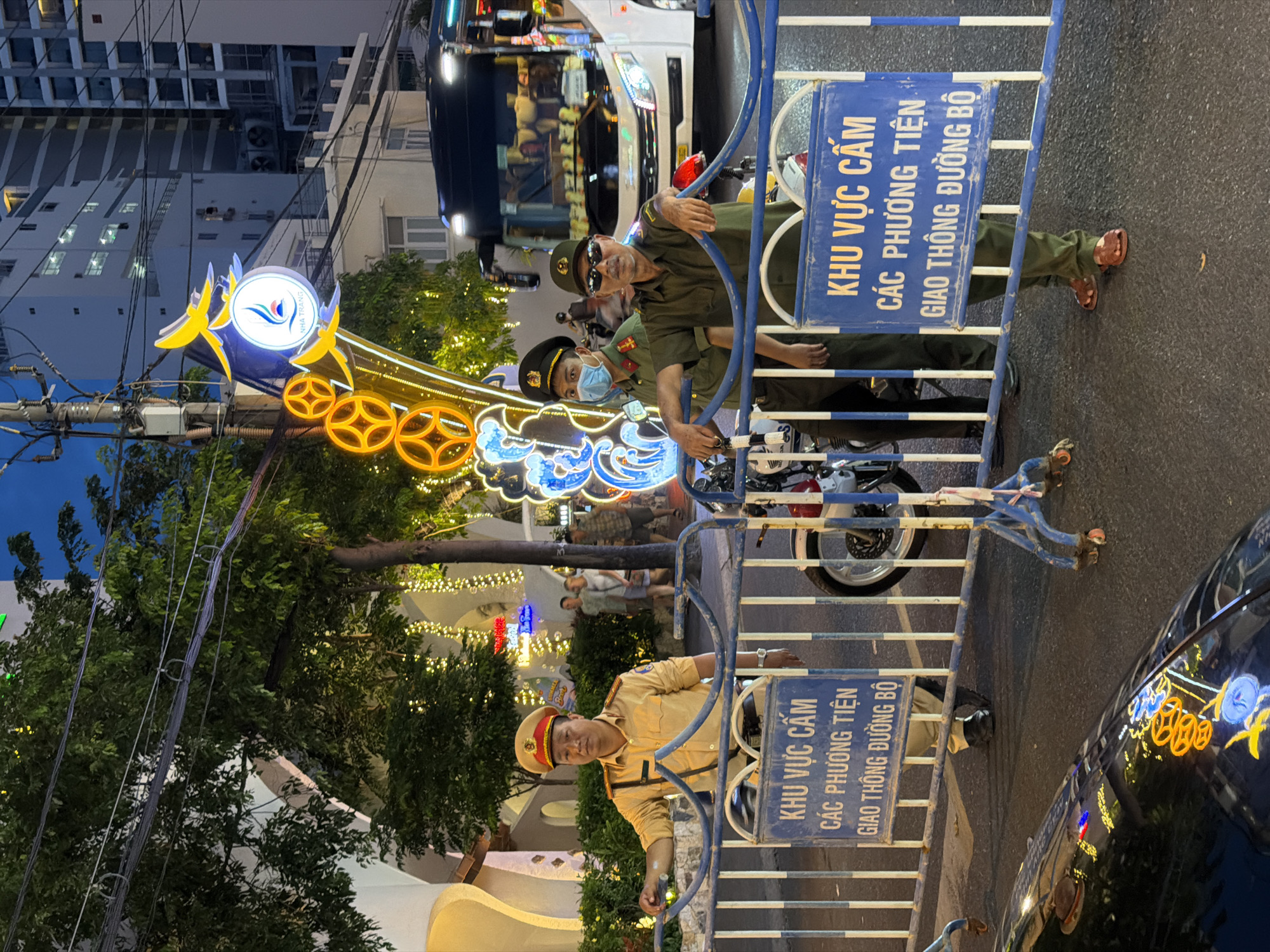 The image shows a nighttime scene in Nha Trang Ward, Vietnam, featuring two Vietnamese police officers, a decorative sign with the words "NHA TRANG", and barricades.
One officer is in an olive-green uniform and sunglasses, another is in a brown uniform, and a third officer, also in an olive-green uniform, has a medical mask on. They are leaning over a blue-and-white metal barricade, which has signs in Vietnamese. One reads: "KHU VỰC CẤM" (RESTRICTED AREA) and "CÁC PHƯƠNG TIỆN GIAO THÔNG ĐƯỜNG BỘ" (ROAD TRAFFIC VEHICLES). A motorcycle is visible behind the barricade.
To the left, there is a decorative sign with the name "NHA TRANG", illuminated with blue and yellow lights, and other lighted decorations. The lights from trees and buildings add to the illumination. In the background, there are buildings, including a tall white apartment building. A black car is visible on the road surface to the right, reflecting some of the light decorations.