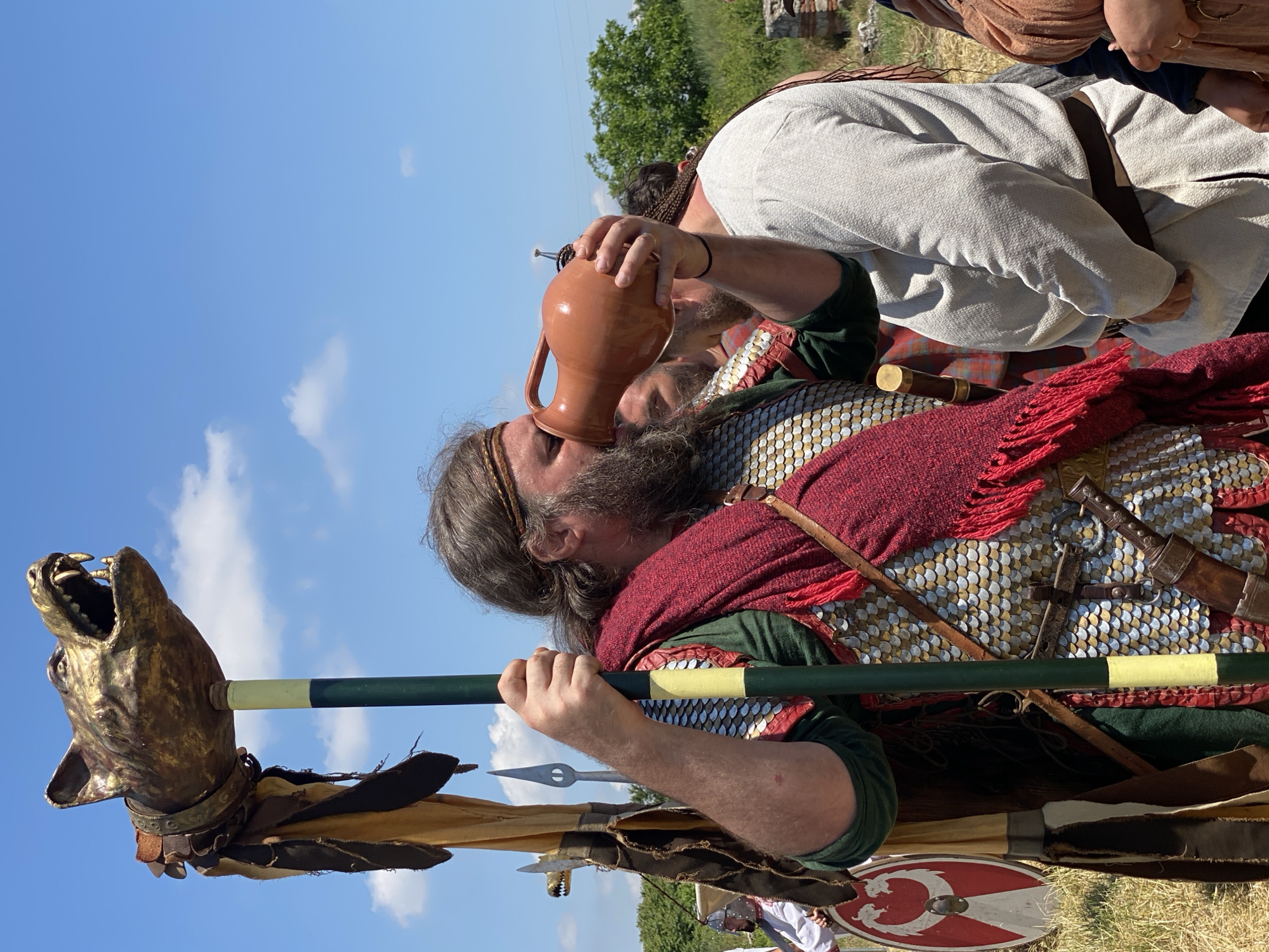 This outdoor image depicts a historical reenactment event in Gigen, Bulgaria, under a clear blue sky with scattered white cumulus clouds. The central figure is a robust man with a long, dark beard and hair secured by a headband. He wears a dark green tunic beneath scale armor, which features a pattern of silver-colored and red scales, and a prominent dark red fringed woolen shawl draped over his left shoulder. His left hand grips a tall, dark green pole with a yellow band, which is crowned by a large, golden-colored, snarling wolf head effigy, possibly a Dacian Draco, from which hang dark leather straps and fabric. With his right hand, he raises a reddish-brown, unglazed clay pitcher to his mouth and drinks.
Behind and to the right of the central figure, a man with braided hair and a light-colored, belted tunic is visible, looking away from the camera. Another bearded man is partially visible behind the central figure's right arm. On the far left, a figure in a white tunic and helmet is partially seen, holding a round shield patterned in red and white, possibly depicting a dragon or serpent. In the background, to the left of the central figure, a smaller, darker wolf head effigy on a pole and a tall, silver-colored spearhead are visible against the sky. The immediate ground is covered in dry grass, while the mid-ground includes green trees and bushes, and faint outlines of stone ruins are discernible on the right horizon.