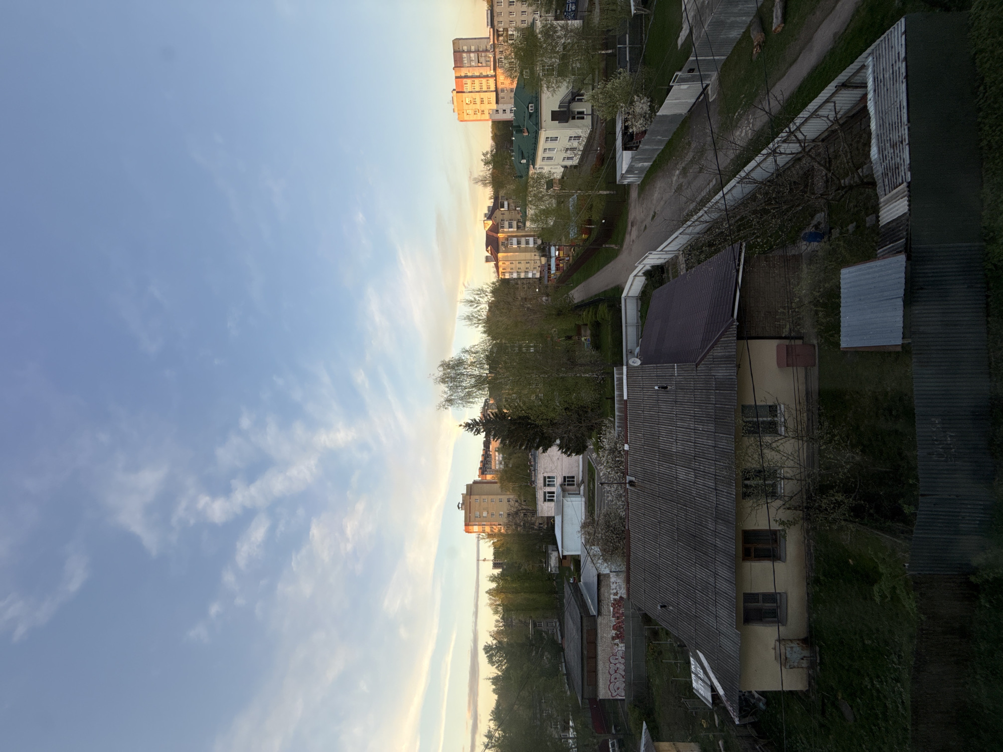 The image presents an elevated perspective of a residential area, likely taken from an upper-floor window or balcony. The scene is divided between a daytime sky with scattered clouds on the left and an urban landscape on the right. The buildings appear to be a mix of low-rise houses and taller apartment blocks, indicating a densely populated area. A road or pathway runs through the center of the view, lined with trees and shrubbery. The buildings closest to the camera display weathered exteriors and varied roof types. The perspective and architectural style suggest the scene is indeed in the Rusanovo area of Russia. No people are visible in the image.