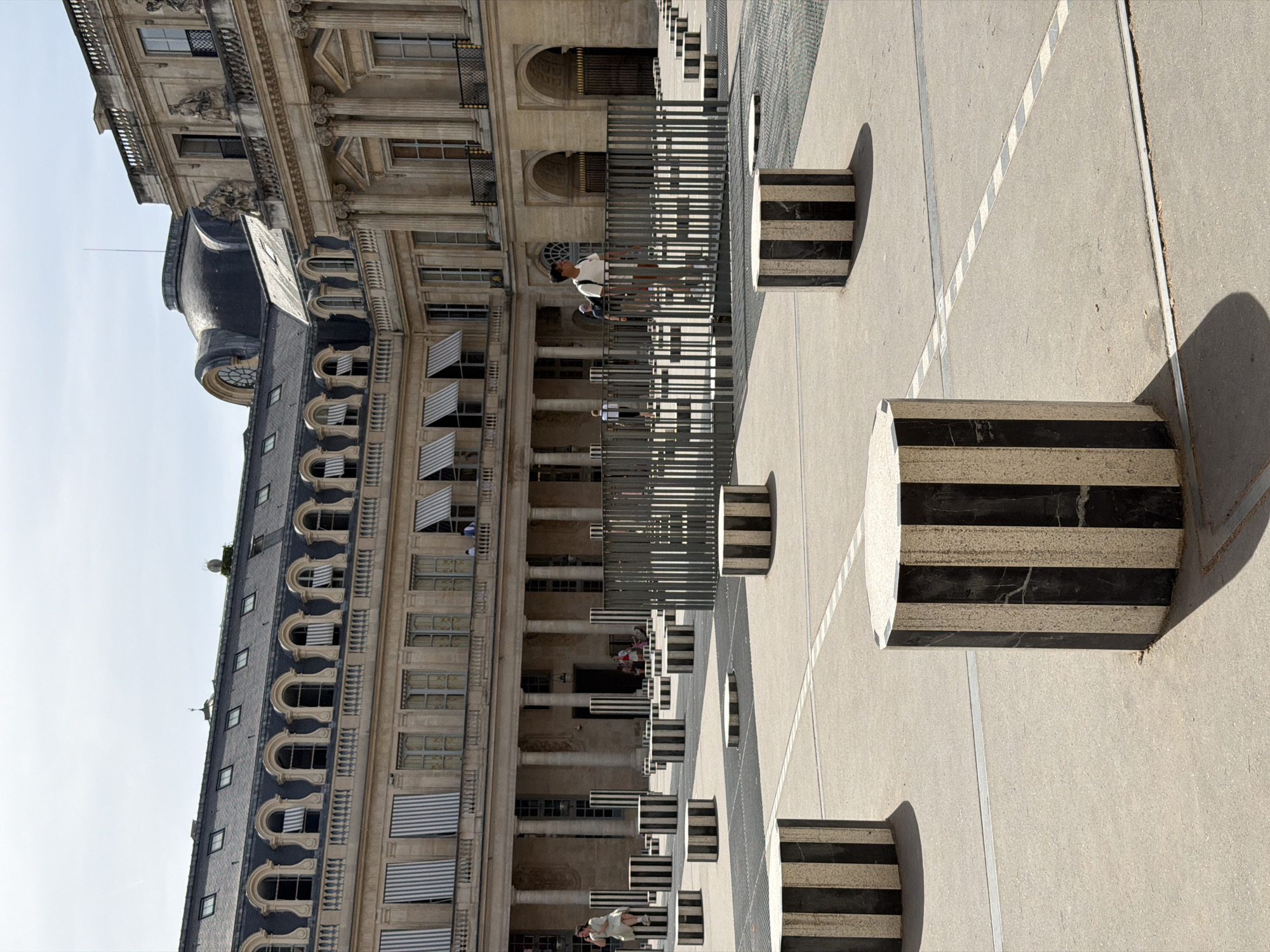 The image captures a sunny day at the Palais-Royal in Paris, France. In the foreground, several large, striped cylindrical columns made of black and white marble stand on a paved courtyard. These are known as the Buren's Columns. Behind the columns, a metal fence with vertical bars runs across the scene. Further back, people are seen walking and interacting within the courtyard. The background features the ornate architecture of the Palais-Royal, with its grand facade, arched windows, and classical columns. The sky is clear and blue.