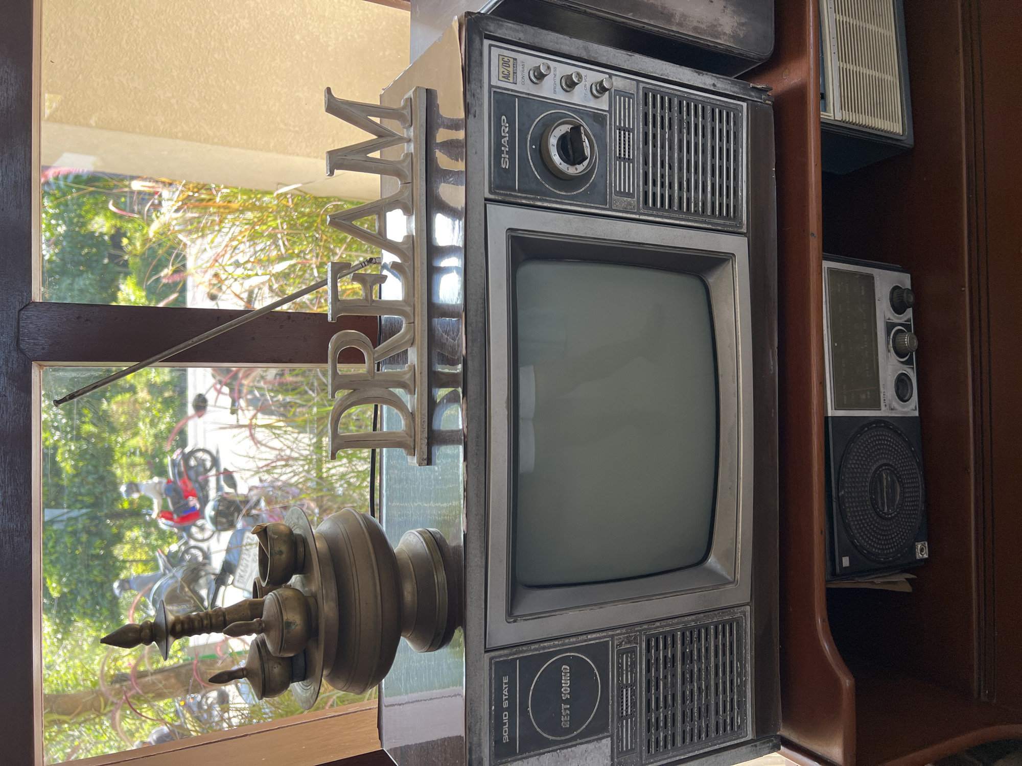In the city of Ubud District, Indonesia, a vintage television set is positioned on a wooden surface. The television screen is dark, and the front of the set displays various buttons and labels, including "SOLID STATE," "BEST SOUND," and "SHARP." To the right of the screen, controls for contrast and brightness are visible. Above the television, a decorative metal word "DREAM" is placed. To the left of the "DREAM" decoration, a traditional brass oil lamp with intricate carvings and small bowls sits. Behind the objects, a window reveals a sunlit outdoor scene with greenery, plants, and people riding motorcycles on a street. Below the television, another shelf holds two smaller electronic devices, one appearing to be a radio. The overall scene suggests a display of vintage items, possibly in a shop or a home setting, with a view of a bustling street outside.