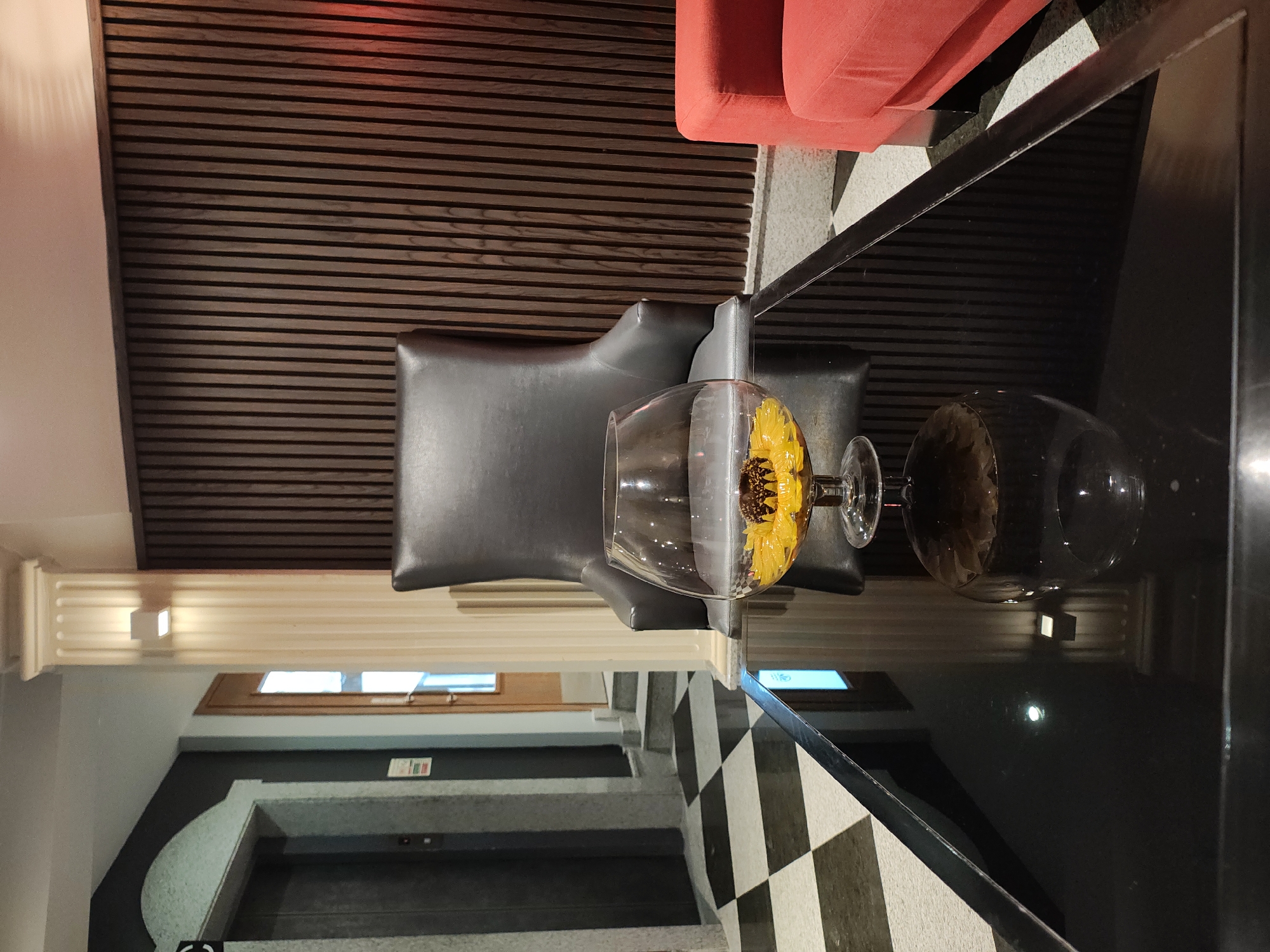 The image depicts an indoor scene, likely a hotel lobby or lounge, with a prominent glass brandy snifter containing yellow flowers and a dark center, placed on a black glass table. To the right of the table is a tall, dark leather armchair, and further to the right, a red sofa. The background features a wall with vertical wooden slats, and to the left, a checkered floor extends towards an elevator and a doorway. The overall impression is one of modern, sophisticated interior design. The image does not provide any explicit visual cues to suggest its location in Manikata, Malta.