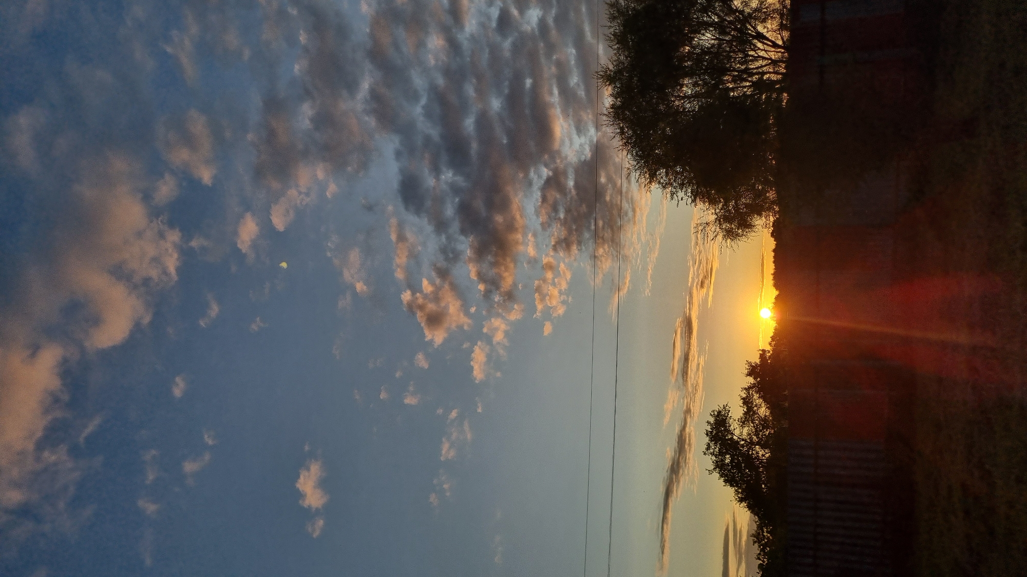 A vertical, low-angle shot captures a sunset over a rural area in Vysokoe, Russia. The sky is a gradient of blues and oranges, with scattered clouds tinged pink and gray by the setting sun. A sliver of the moon is visible in the upper right quadrant of the sky. In the mid-ground, a fence runs horizontally across the frame, silhouetted against the bright horizon. Trees and foliage are also visible in silhouette on either side of the fence. The foreground consists of shadowed grass. Power lines stretch across the sky in the upper mid-ground. The sun itself is a bright orb low on the horizon, casting long rays of light and lens flare downwards. No people are visible in the scene.
